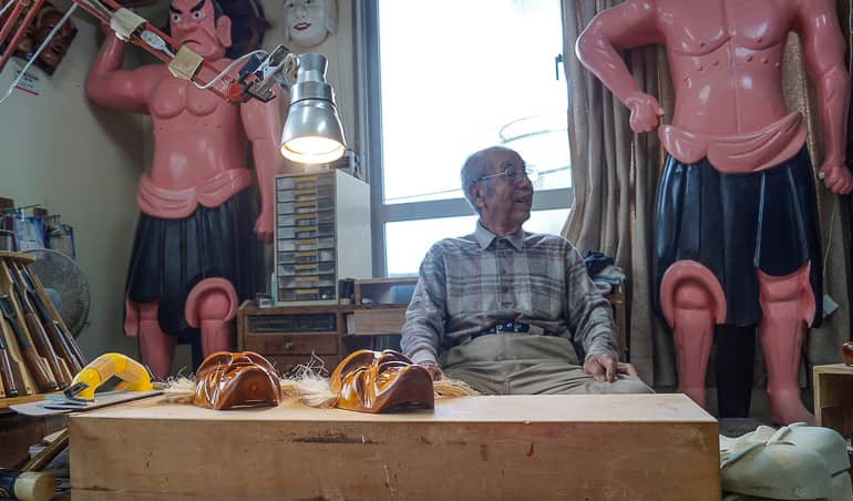 Mr. Taba lachend an seinem Tisch, vor ihm fertige und unfertige Masken.