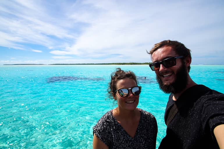 Die beiden Blogger Anne und Clemens lächeln von einem Vaka auf dem hellblauen Meer der Cook Insel Aitutaki strahlend in die Kamera.