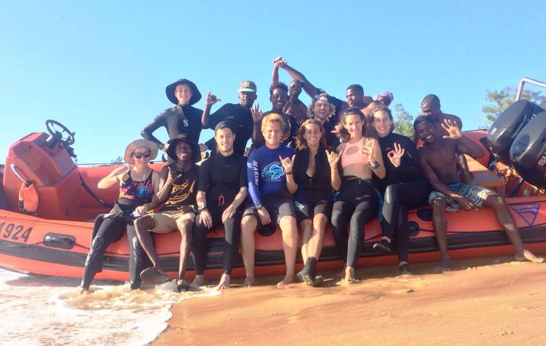 In einem Surfcamp sitzt eine Gruppe junger Menschen zusammen auf einem Boot; schnell neue Leute beim Solo Travel kennenlernen klappt in Camps besonders gut.