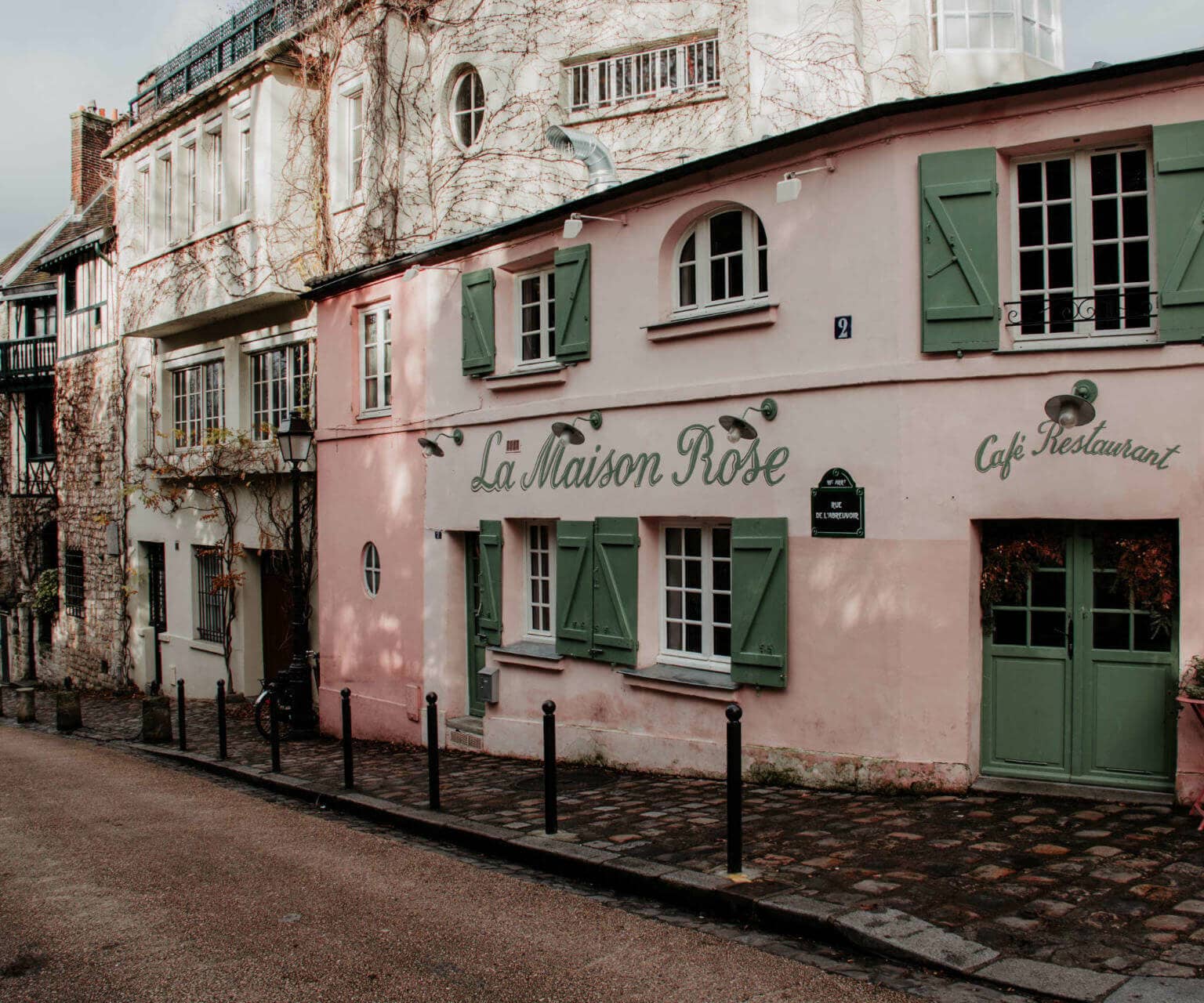 La Maison Rose ist ein Pariser Café und Restaurant in einer kleinen Straße mit rosa Fassade und grünen Fensterläden.