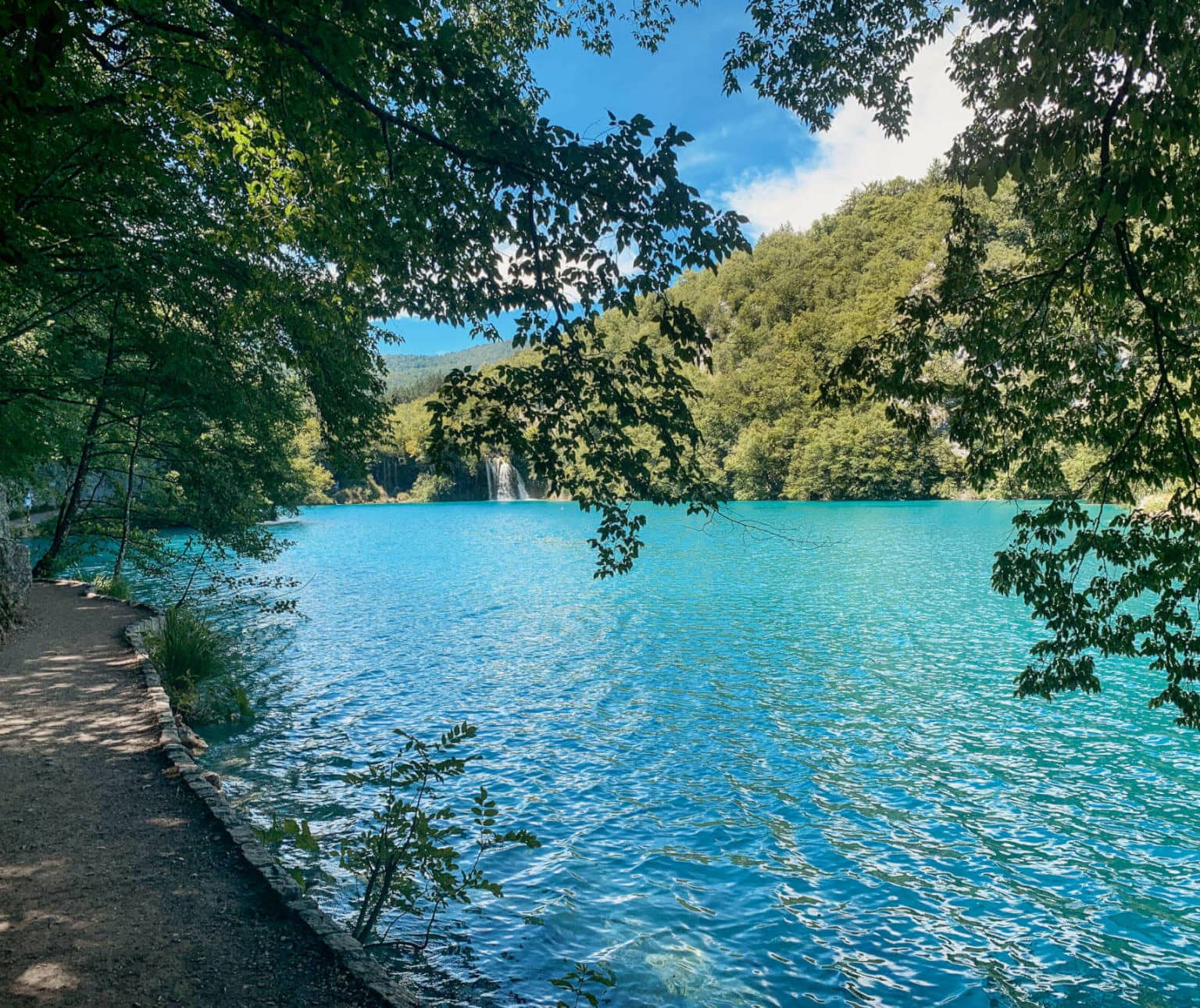 Neben einem glasklaren, türkisfarbenen See im Nationalpark Plitvicer Seen in Kroatien führt ein unter Bäumen liegender Wanderweg entlang.