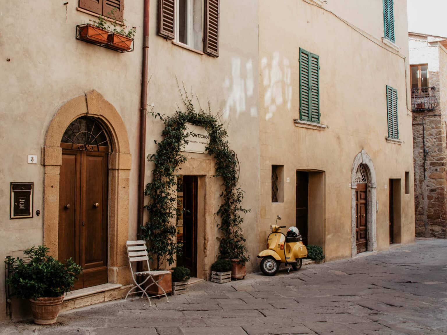 Eine gelbe Vespa steht an ein Sandsteinhaus gelehnt, die Hauswand zieren Fenster mit grünen Fensterläden, schwere Holztüren und ein Pflanzenkranz um die Eingangstür gebunden.