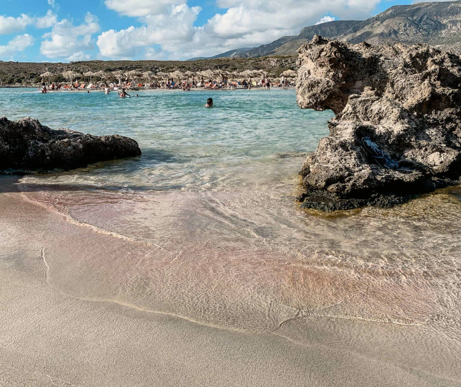 Sandstrand in Elafonissi auf Kreta gesäumt von Felsen, im Hintergrund liegen Besucher unter Strohsonnenschirmen oder schwimmen im Meer.