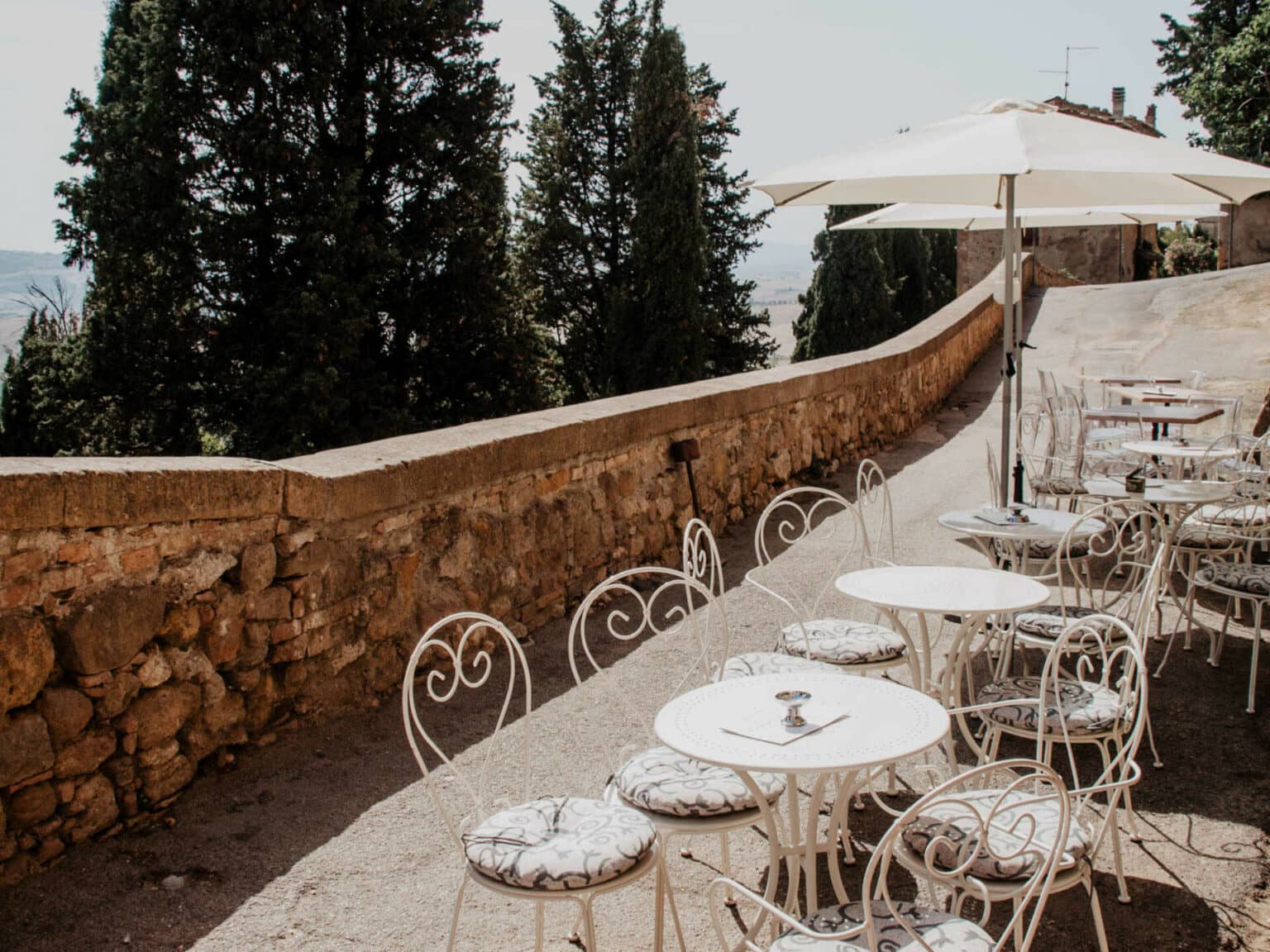 Die Sonnen-Terrasse einer Bar lädt mit weißen Stühlen in verschnörkeltem Design und Sonnenschirme zu einer Aussicht auf die bezaubernde toskanische Landschaft mit ihren Zypressenbäumen ein.