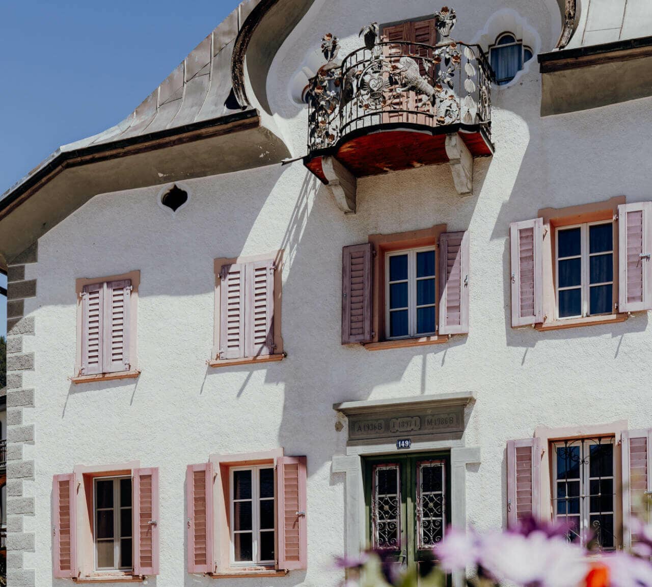 Ein weißes Haus im Jugendstil mit rosa Fensterläden und einem verschnörkelten Balkon im zweiten Stock, in Sent, Schweiz.