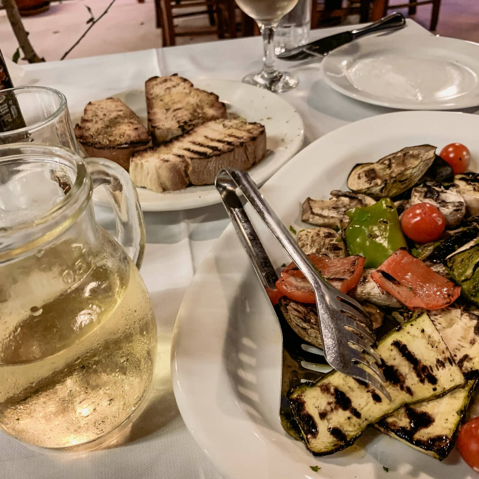 Auf einem Tisch angerichtetes Essen, geröstetes Gemüse, eine Karaffe mit Weißwein und Brot.