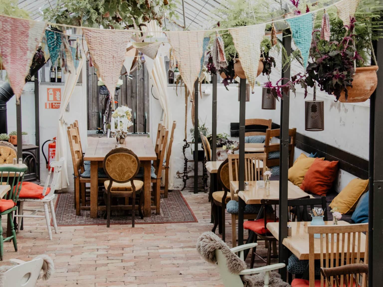 In einem kleinen und gemütlichen Café stehen Holztische und -stühle zusammen, am rechten Rand steht eine Couch mit bunten Dekokissen. Pflanzen und eine Girlande aus Makramee schmücken den Raum.