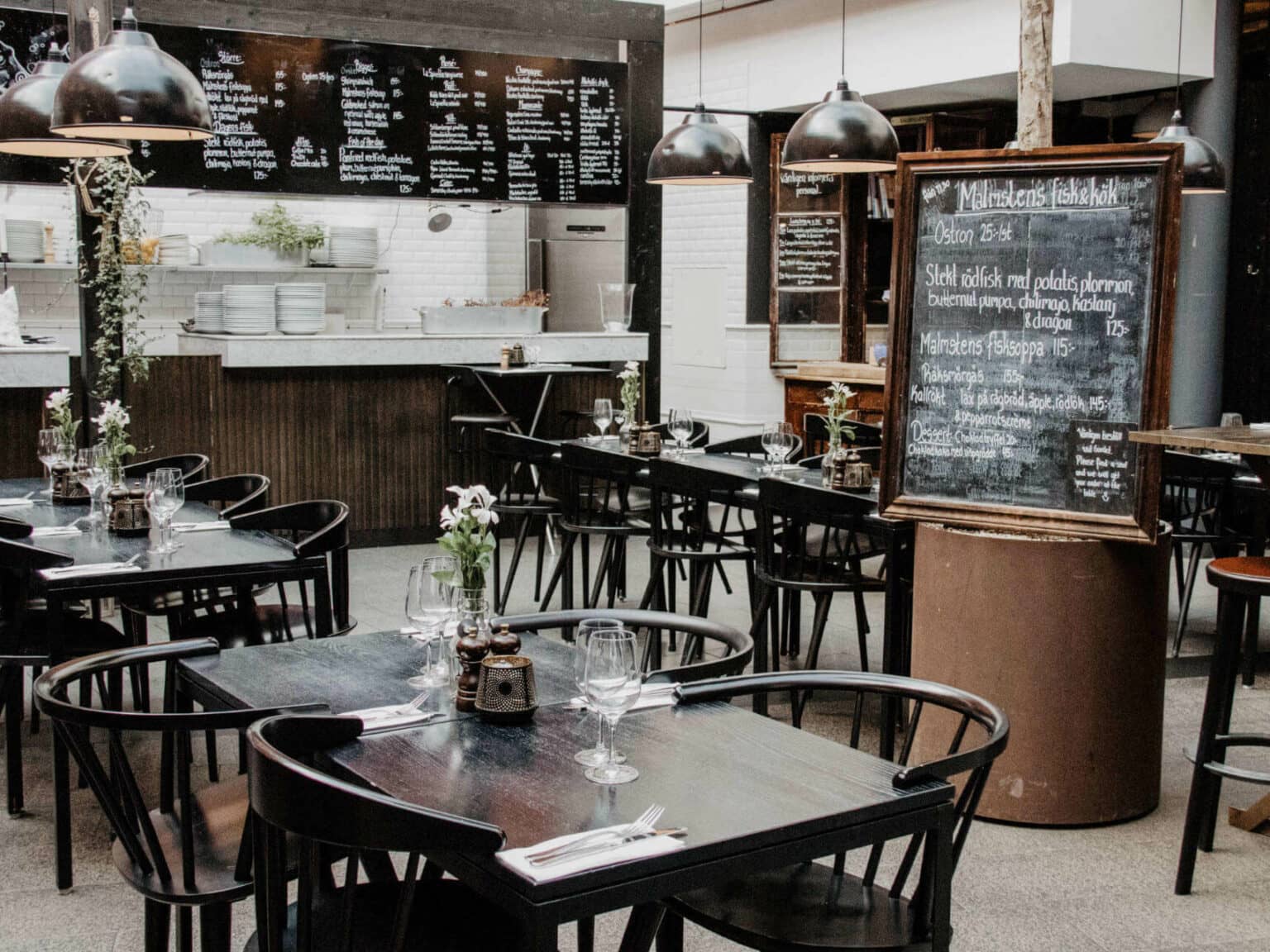 In einer Markthalle stehen mehrere schwarze Stühle und Tische zusammen, auf den Tischen sind rosa Blumen in Vasen angerichtet, von der Decke hängen schwarze Industrieleuchten; Kreidetafeln kündigen Speisen und Getränke an.