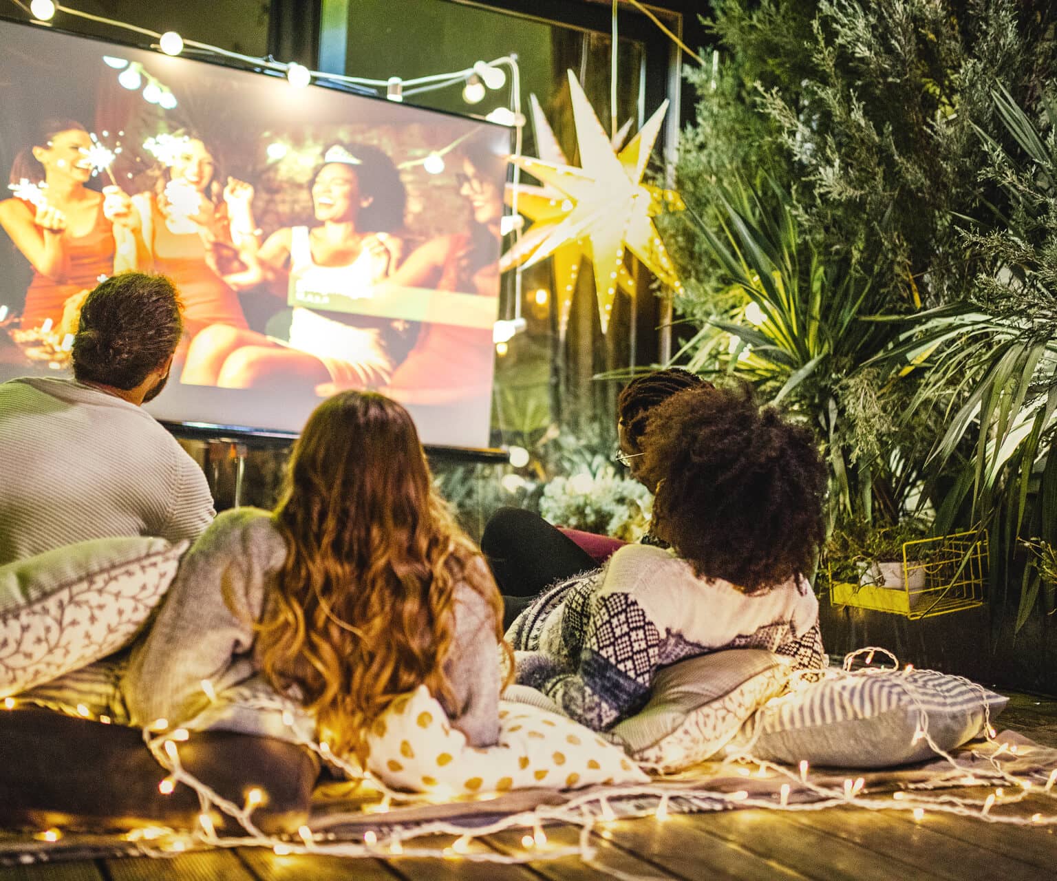 Drei Freunde liegen auf Decken im Hinterhof und schauen einen Film auf einer Leinwand.