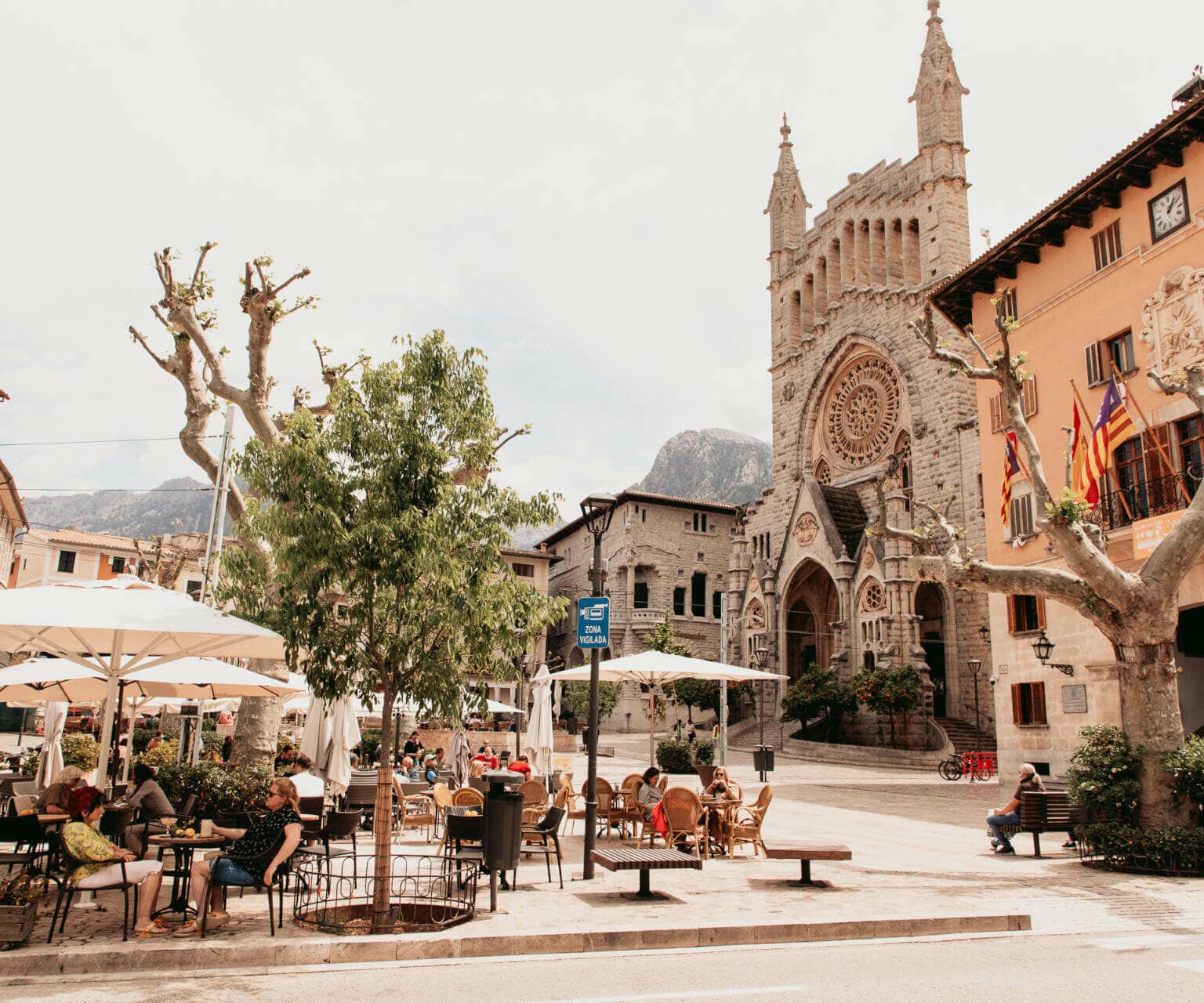 Der belebte Plaça Constitució in Sóller mit Menschen unter Sonnenschirmen und Blick auf die Kirche Sant Bartomeu auf Mallorca.