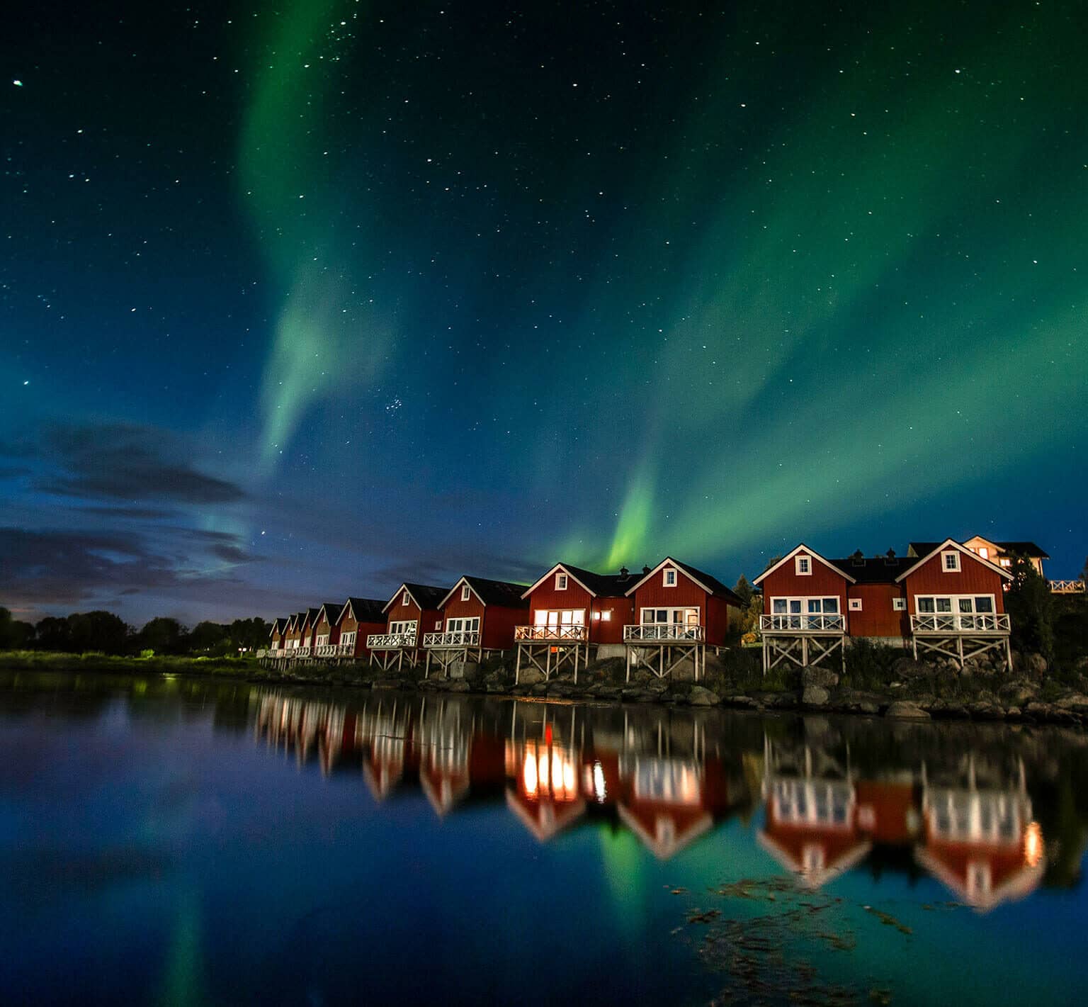 Die Polarlichter scheinen bei Nacht über eine Reihe an roten Hütten am Wasser
