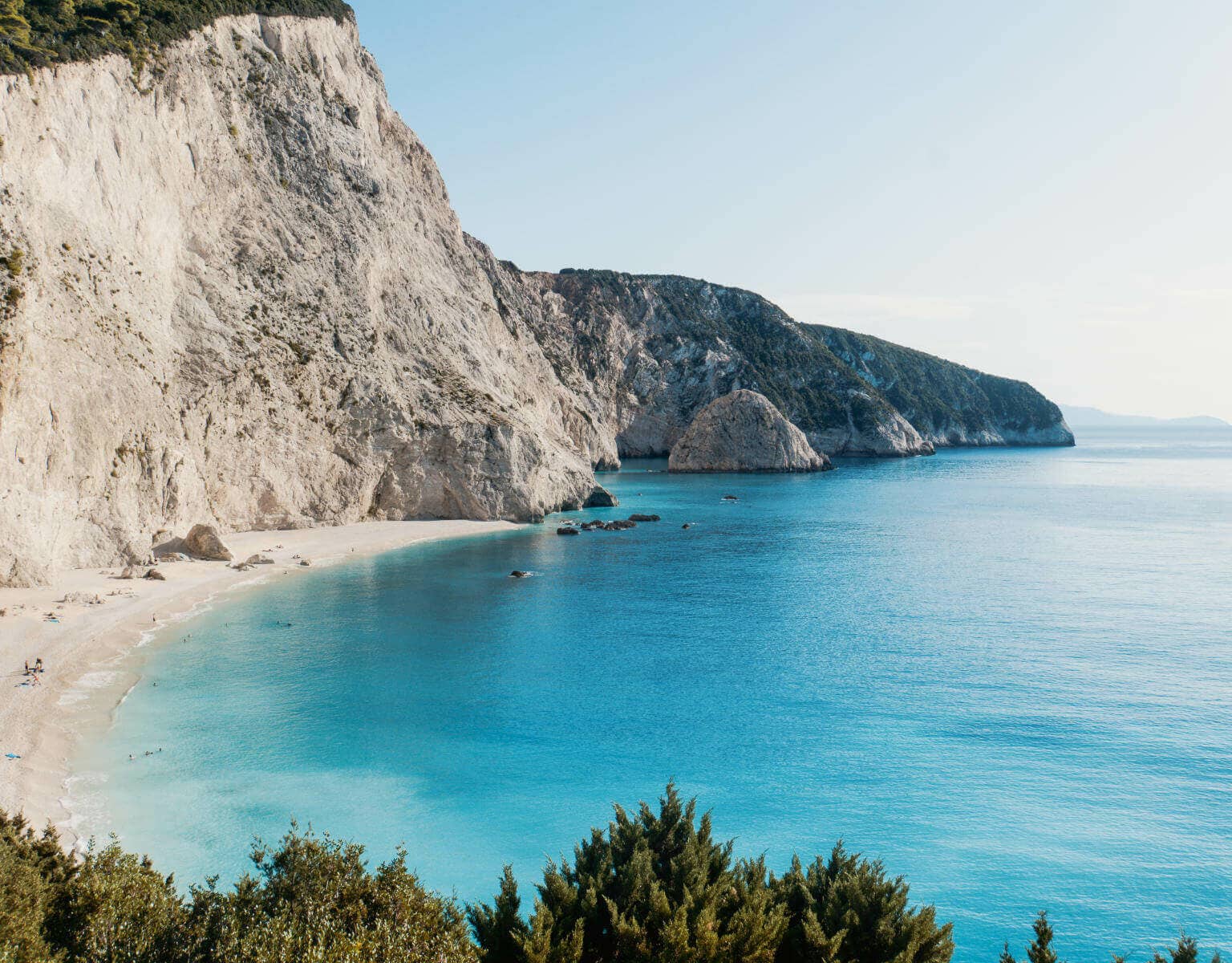 Hohe Wände aus Kalkfelsen umrahmen die traumhafte Bucht von Porto Katsiki auf der griechischen Insel Lefkada. Besucher sonnen sich am feinen Sandstrand, ein paar von ihnen baden in dem türkisblauen Wasser des Ionischen Meers.