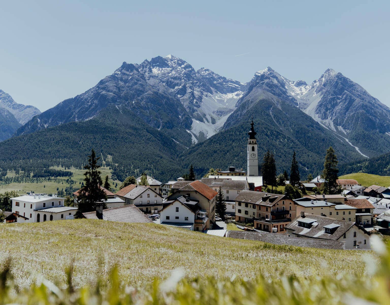 Blick über eine Wiese hinein in ein Dorf mit Kirchturmspitze, dahinter Bergpanorama, in Ftan in der Schweiz.