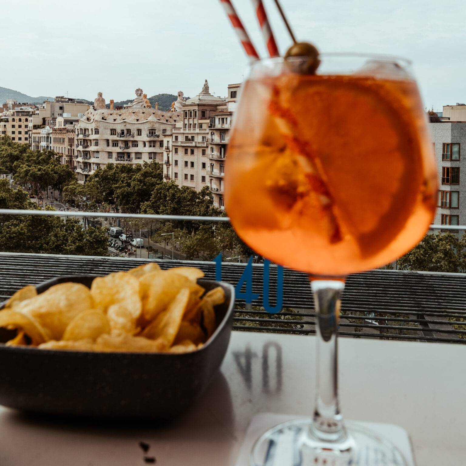 Glas Aperol Spritz und eine Schale Chips stehen auf eine Tisch und man blickt über die Häuser von Barcelona