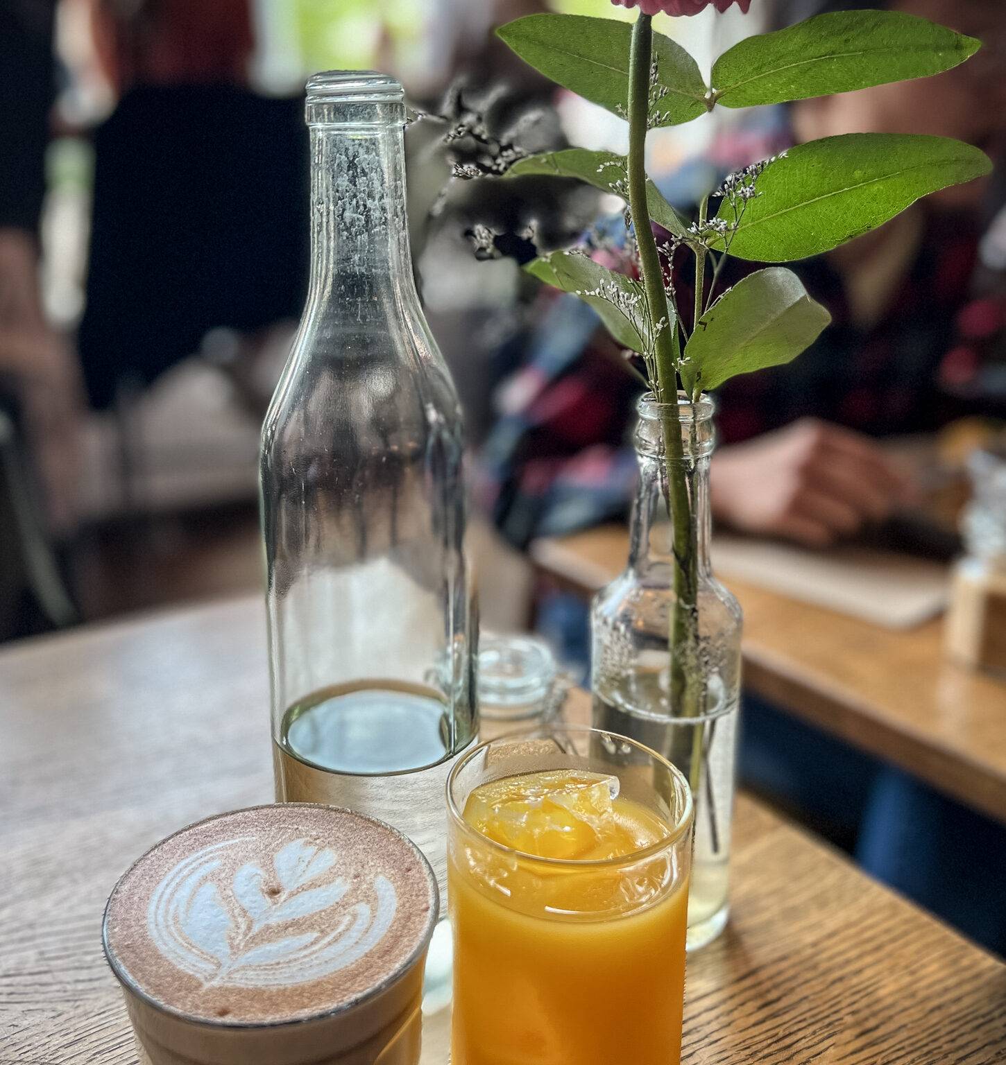 Auf einem Holztisch steht eine Flasche mit Wasser, ein Glas Orangensaft, ein Cappuccino und eine Vase mit einer Blume.