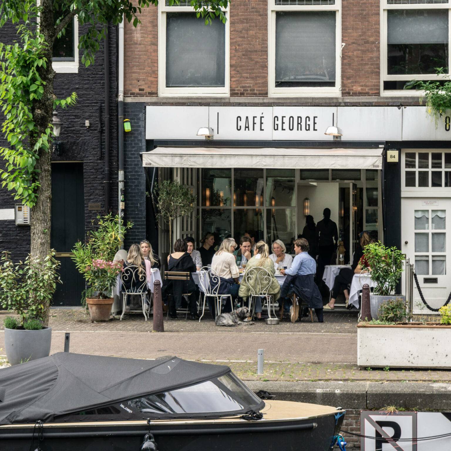 Vor einem Café direkt am Wasser sitzen einige Menschen an Tischen und unterhalten sich.