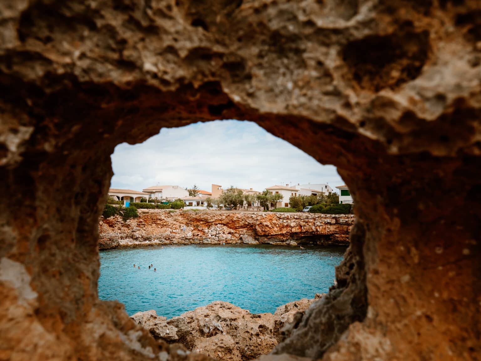 Blick durch ein Lochfenster im Fels auf die Bucht Cala Morlanda mit blauem Meer und felsigen Naturgestein.