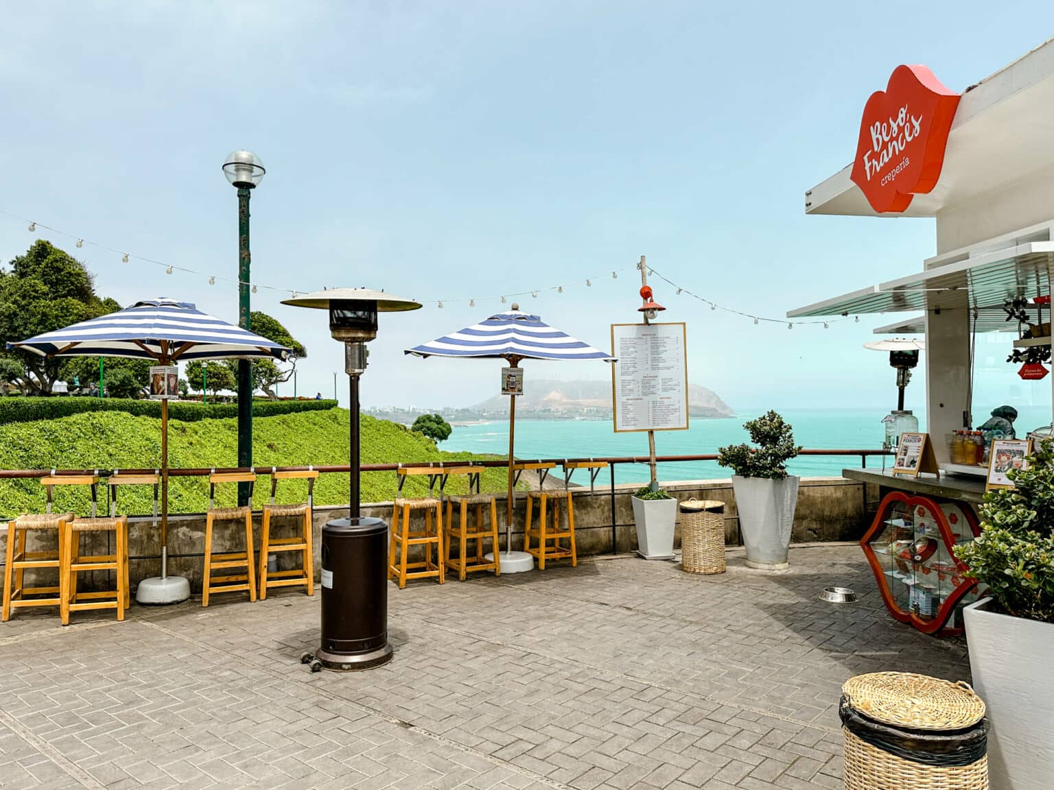 Ein kleines Cafe an der Küstenpromenade von Lima mit blau weißen Schirmen und Blick auf das Meer.