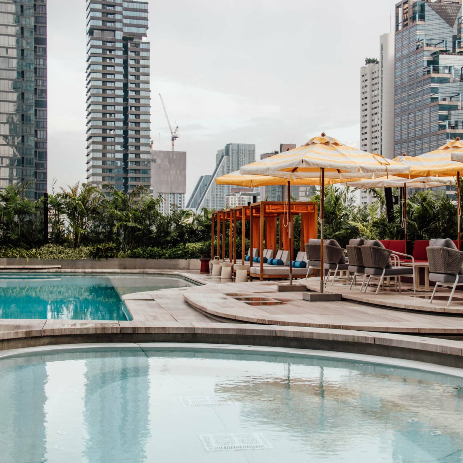 Ein großer Hotelpool im Vordergrund umgeben von Palmen und Hochhäusern inmitten von Bangkok.