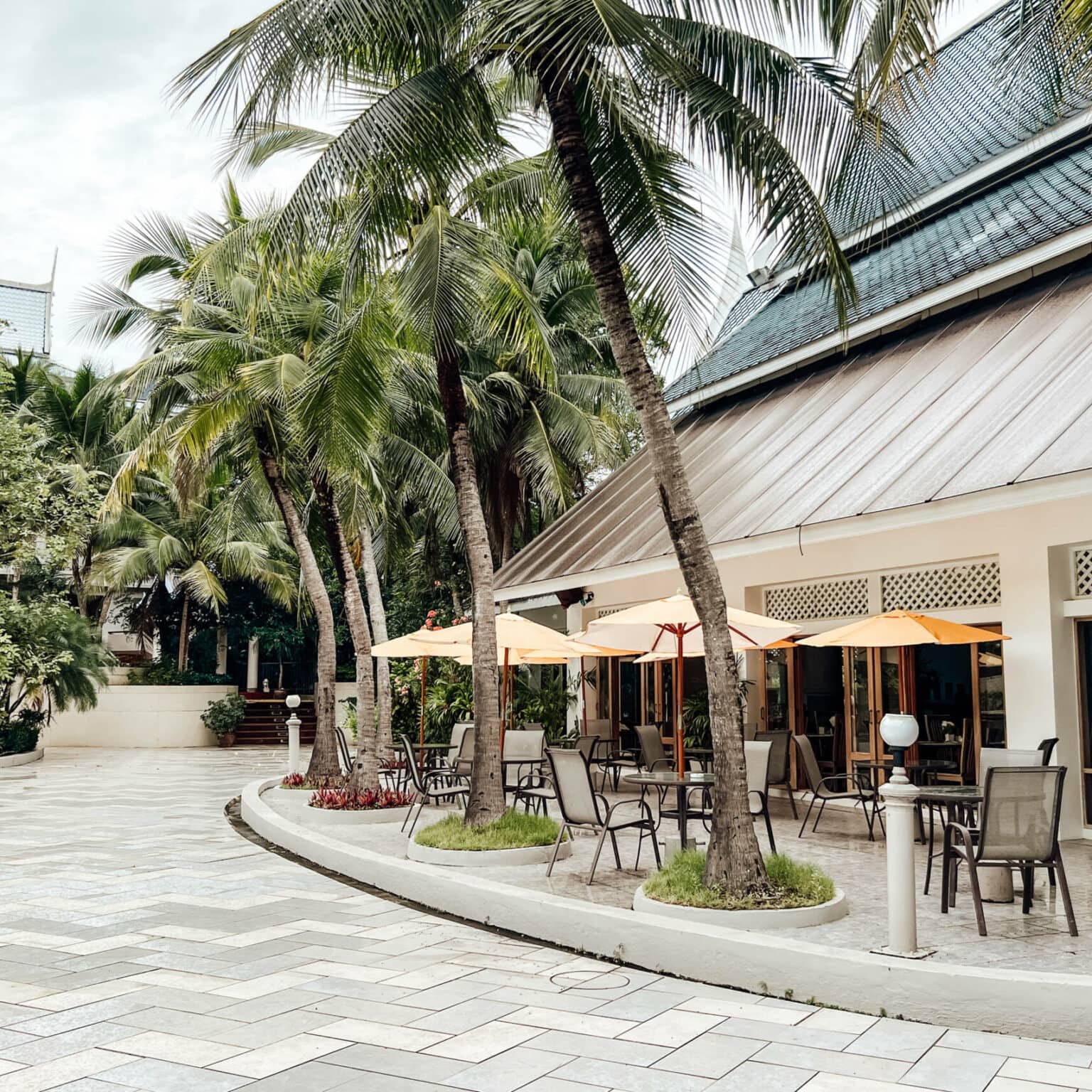 Der palmengesäumte Außenbereich des Chada Thai Village Resort in Krabi mit Tischen, Stühlen und gelben Schirmen.