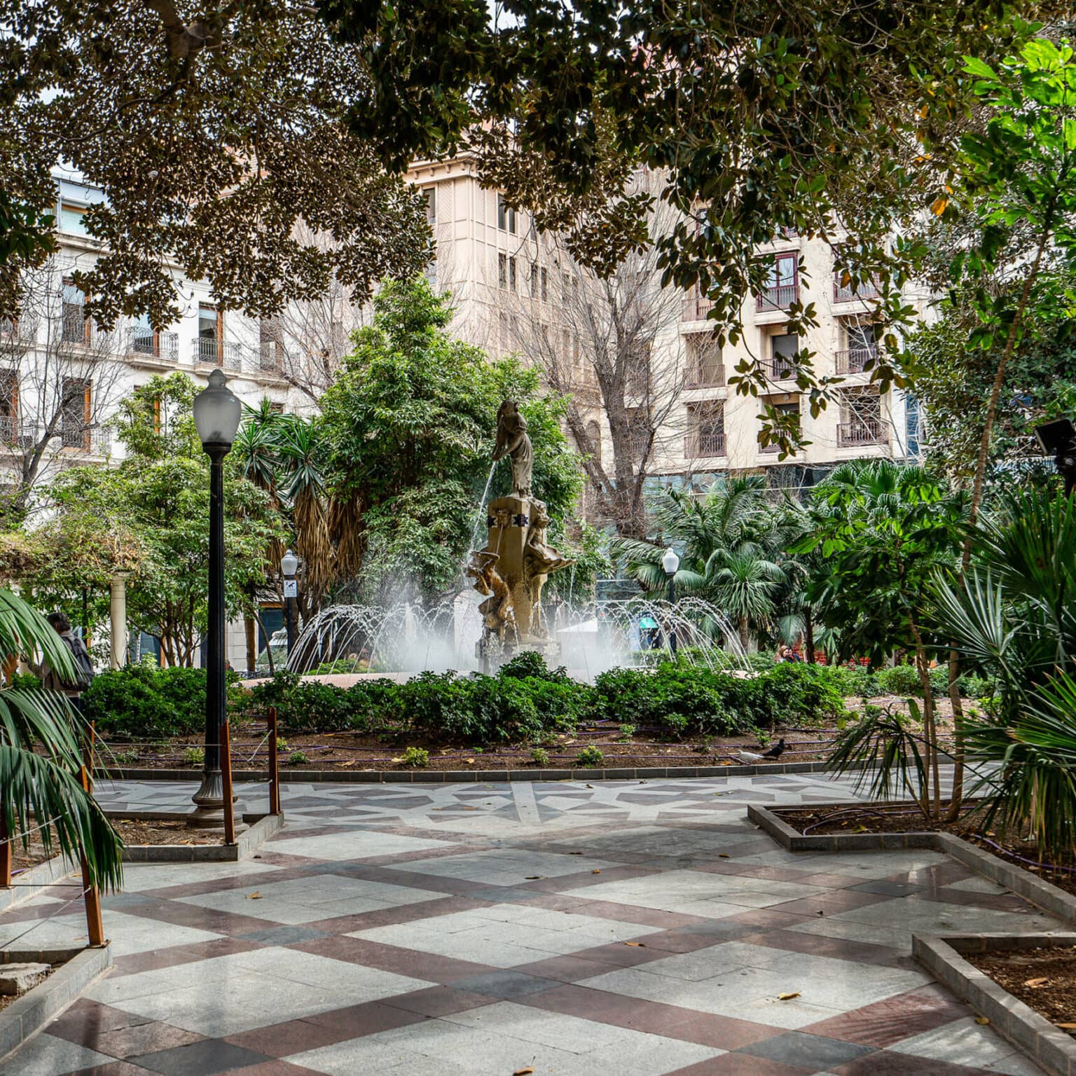 Ein Brunnen inmitten der Altstadt von Alicante zwischen vielen Bäumen und Palmen.