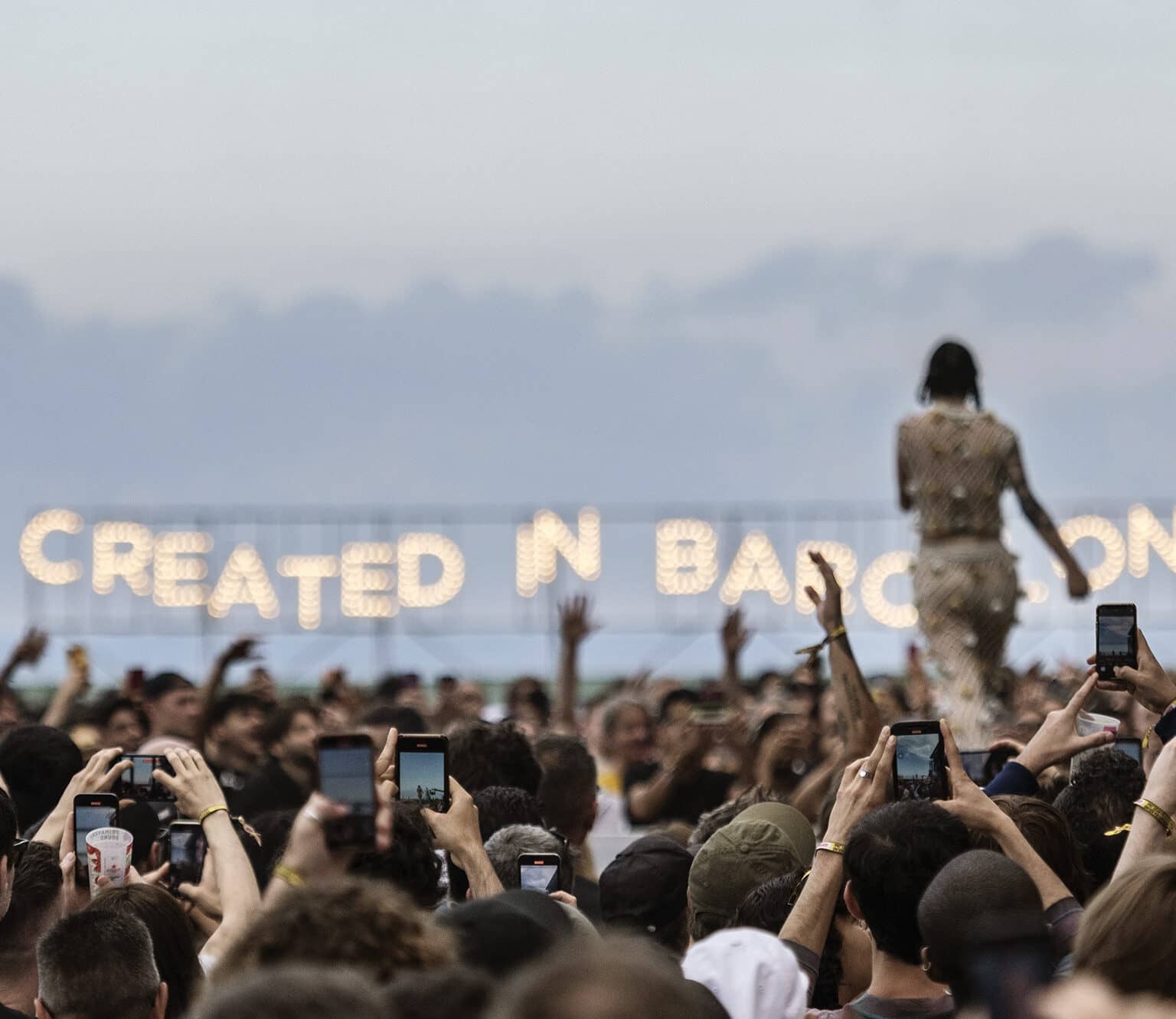 Eine jubelnde Menge vor einer Bühne bei Tag. Einige filmen mit ihren Handys. Im Hintergrund läuft die Künstlerin und und es gibt ein Schild mit der Aufschrift "CREATED IN BARCELONA".