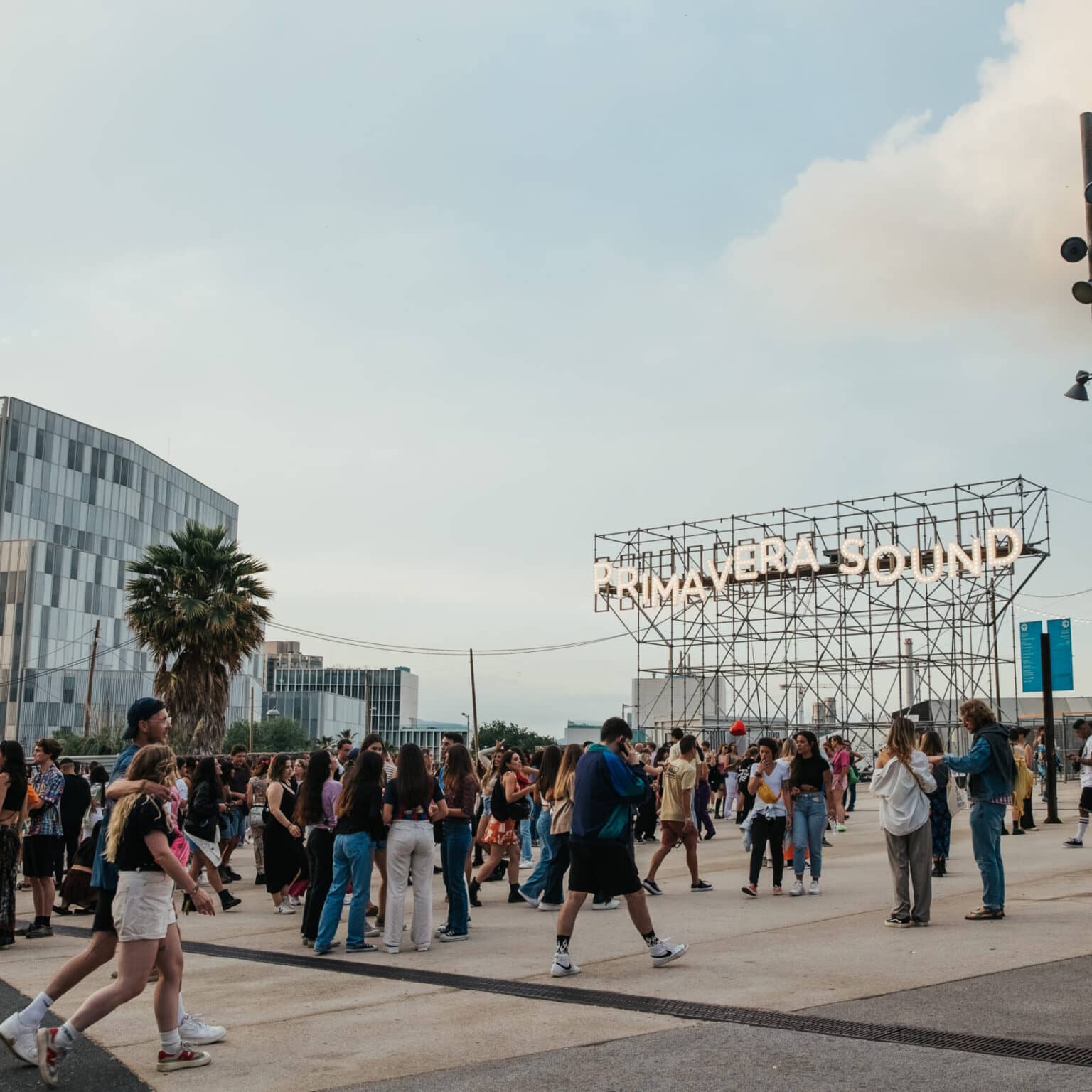 Einige Menschen laufen über einen Platz. Im Hintergrund steht ein Gerüst mit der leuchtenden Aufschrift "PRIMAVERA SOUND".