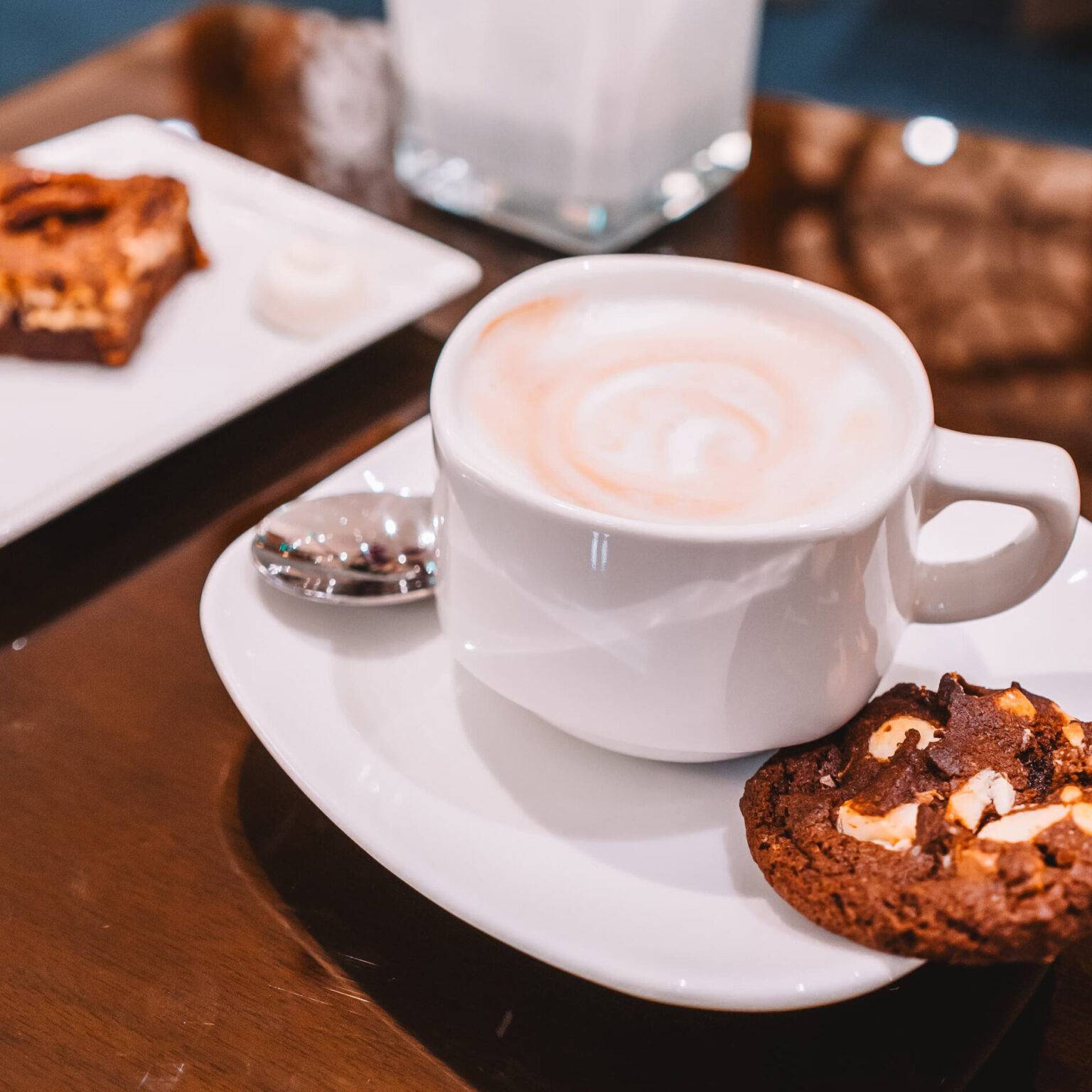 Auf einem Tisch stehen eine Tasse Kaffee mit einem Keks, ein Teller mit einem Stück Kuchen und eine Orchidee.