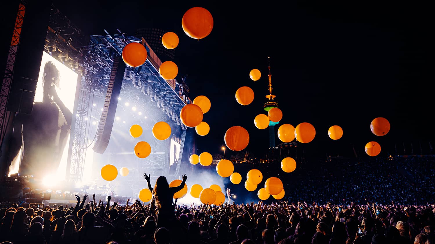Eine jubelnde Menge steht vor der Bühne im Olympia-Stadion. Ein Künstler tritt auf und große orangefarbene Luftballons fliegen über die Menge.