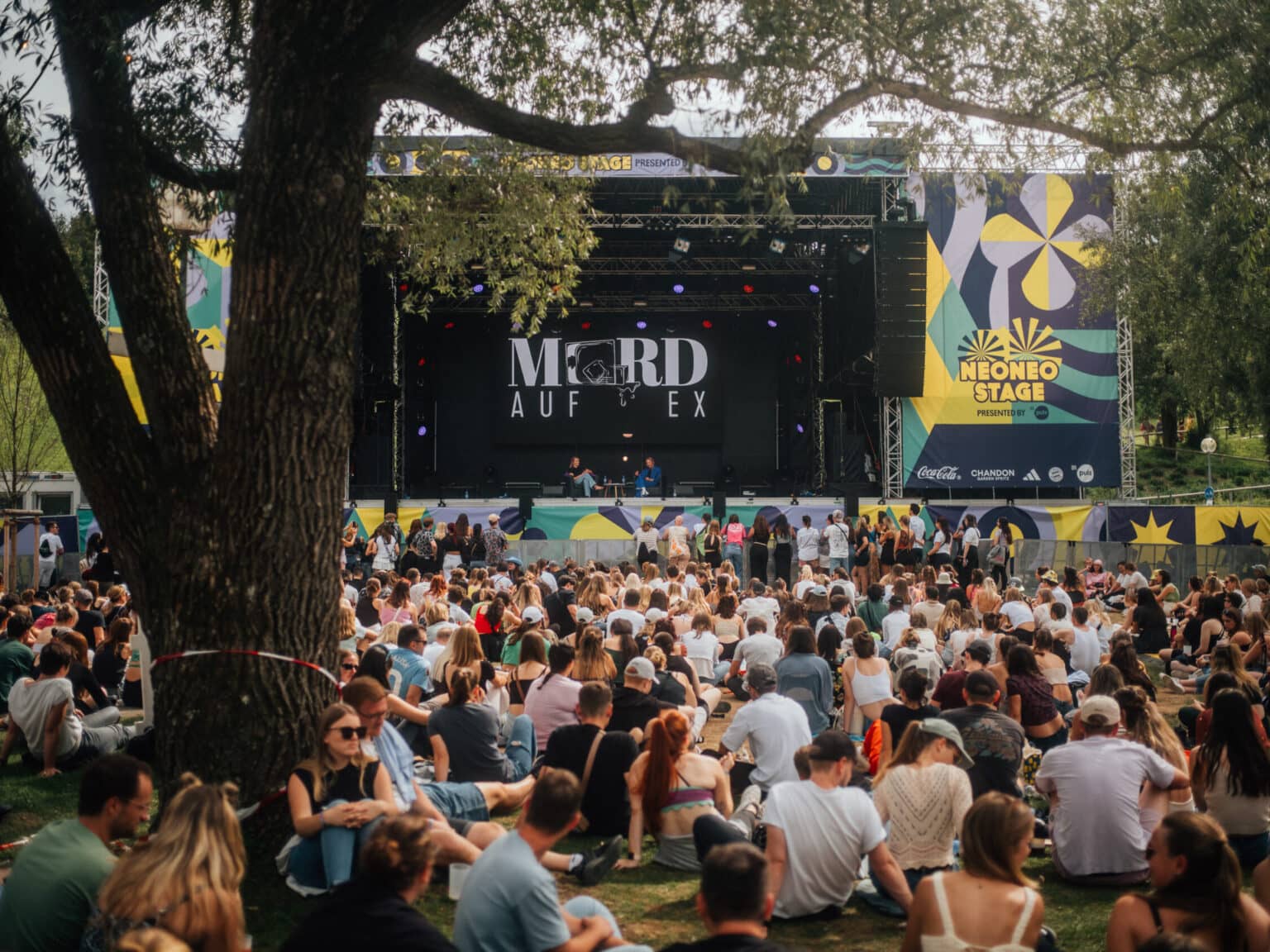 Einige Menschen sitzen auf einer Wiese vor einer Bühne auf der gerade ein Mord auf Ex Live-Podcast stattfindet.