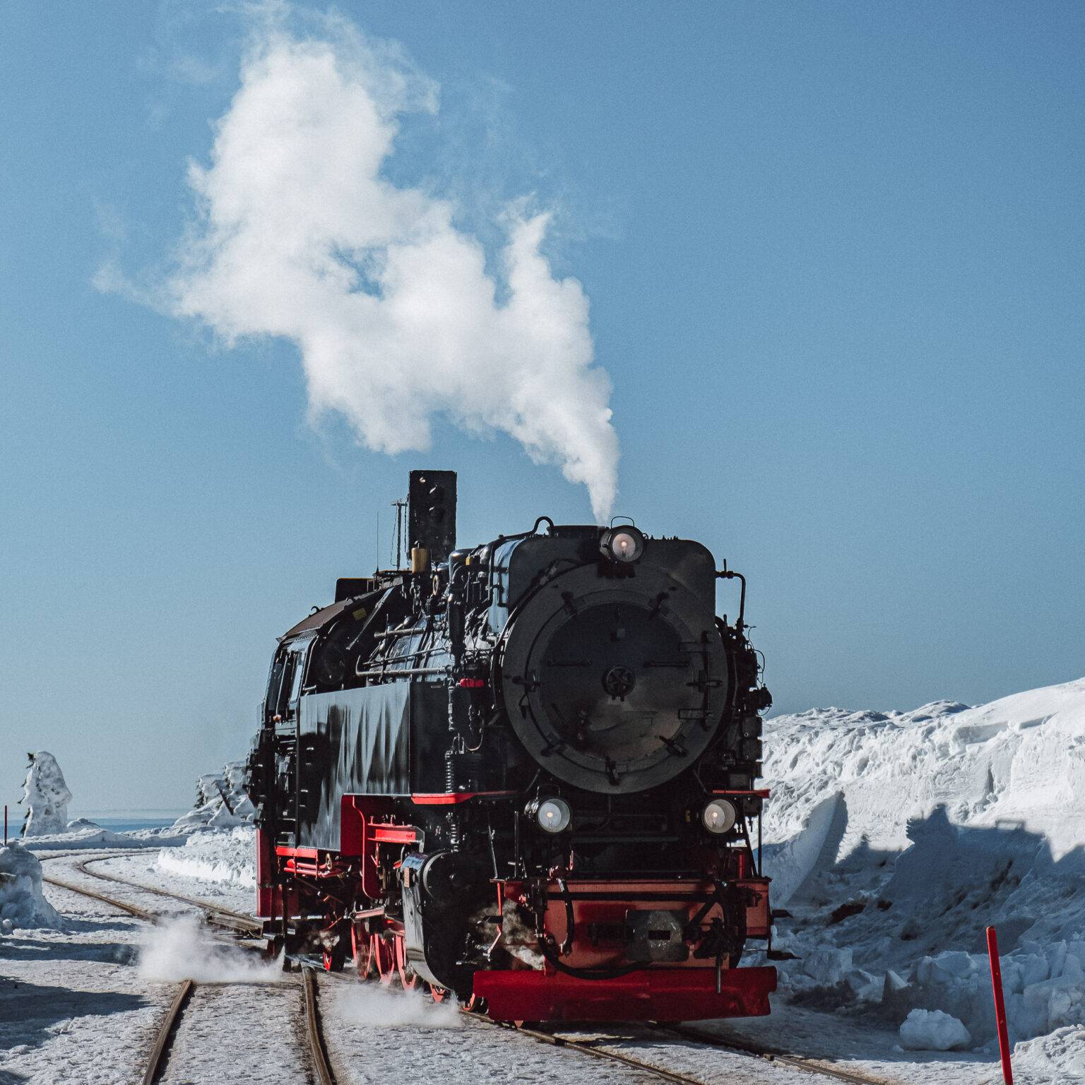 Die Brockenbahn, eine rot-schwarze Lokomotive aus Metall fährt durch eine verschneite Landschaft. Aus ihrem Schornstein steigt Rauch auf.