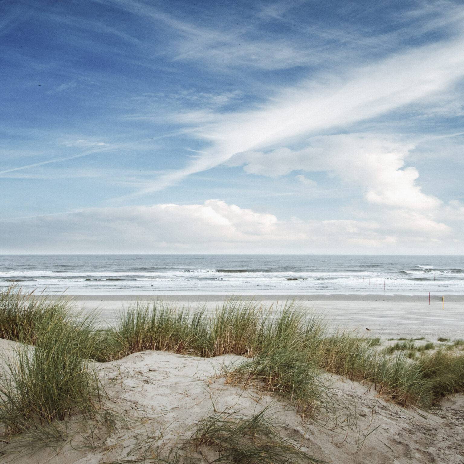 Ein Strand auf Langeoog mit Dünen. Das Meer schlägt Wellen und am blauen Himmel sind vereinzelte weiße Wolken.