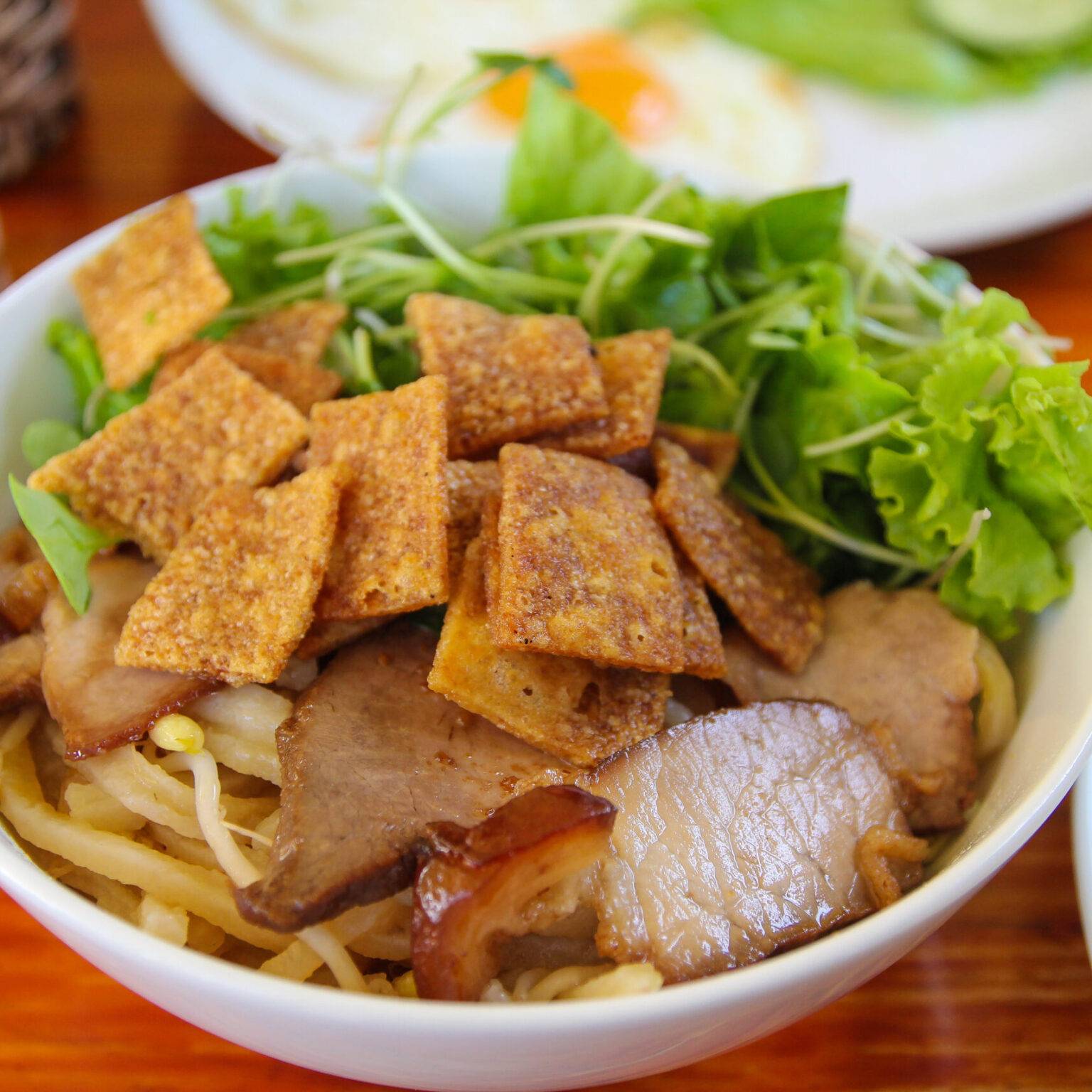 Eine Schüssel mit dem Gericht Cao Lau mit mit Weizennudeln, Schweinefleisch, Salat und Reiscrackern.