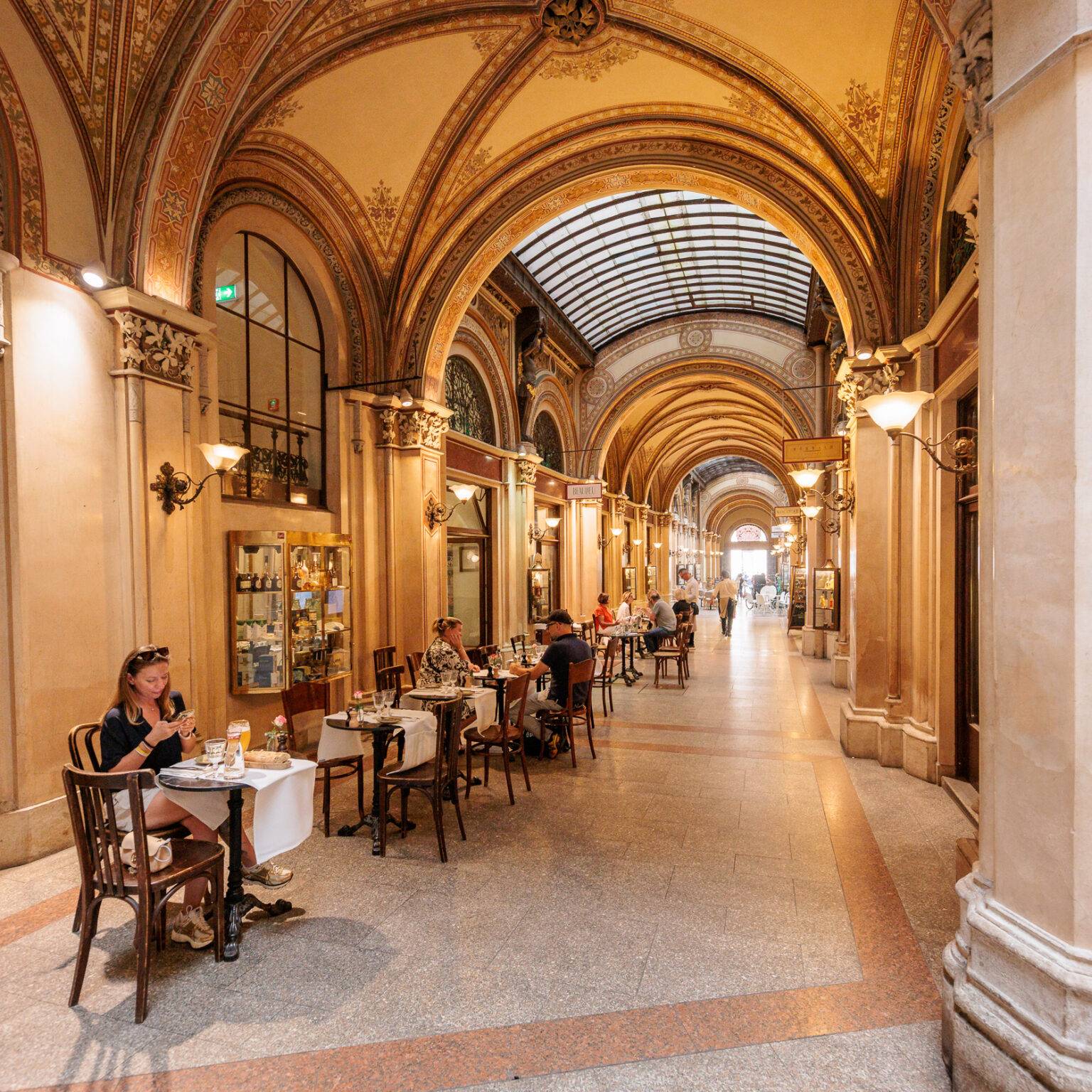 Elegante, beleuchtete Arkade in der Ferstelpassage mit Tischen und Stühlen, die zu einem Restaurant gehören, und einem langen, gewölbten Gang.