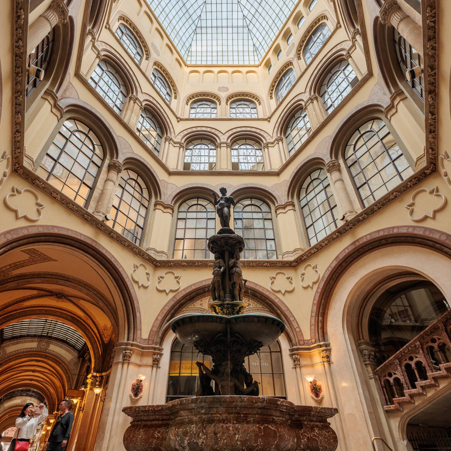 Das Innere der Ferstelpassage mit einer zentralen Statue und einem großen Glasdach, umgeben von Säulen und Bögen.