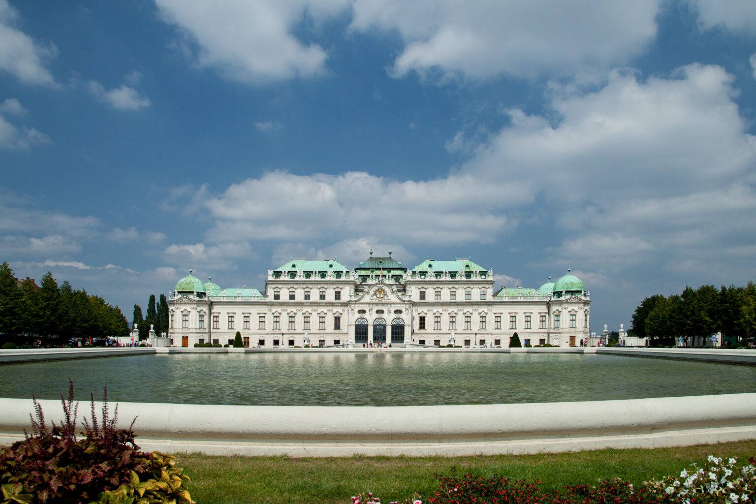 Barockes Schloss Belvedere mit grünen Dächern, umgeben von einem Teich und bunten Blumen im Vordergrund.