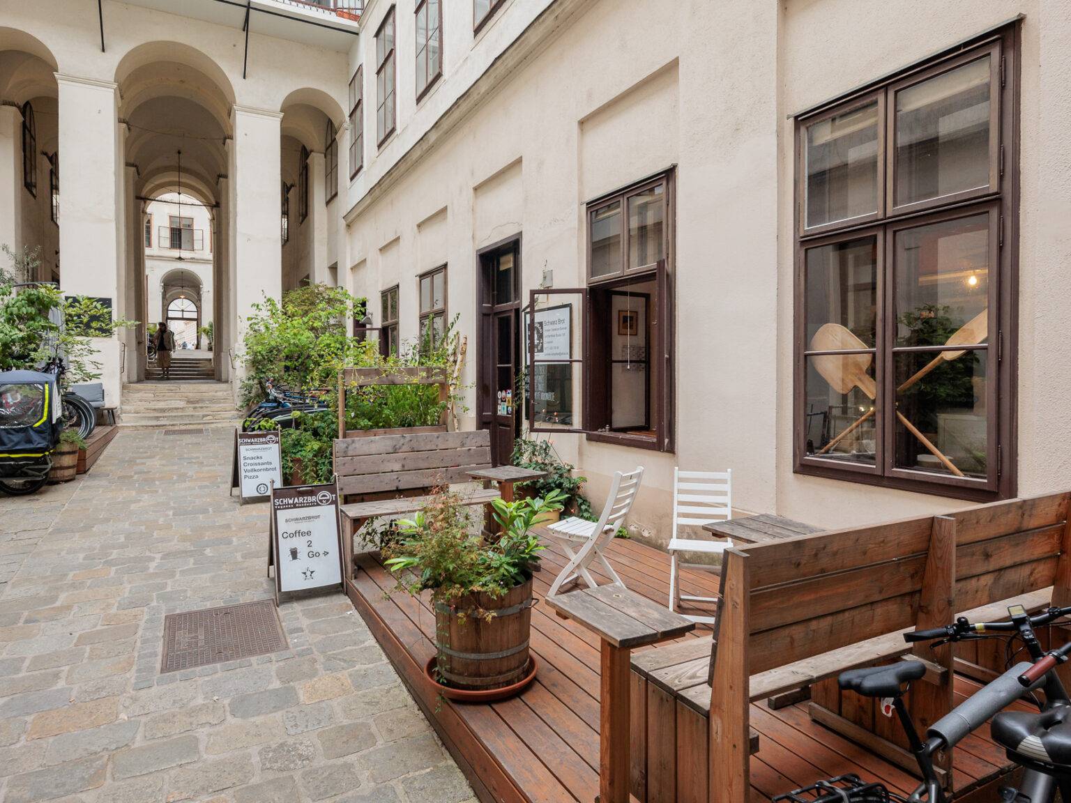 Die hölzerne Terrasse der Bäckerei Schwarzbrot in einem gepflasterten Innenhof mit Sitzgelegenheiten und Pflanzenkübeln.