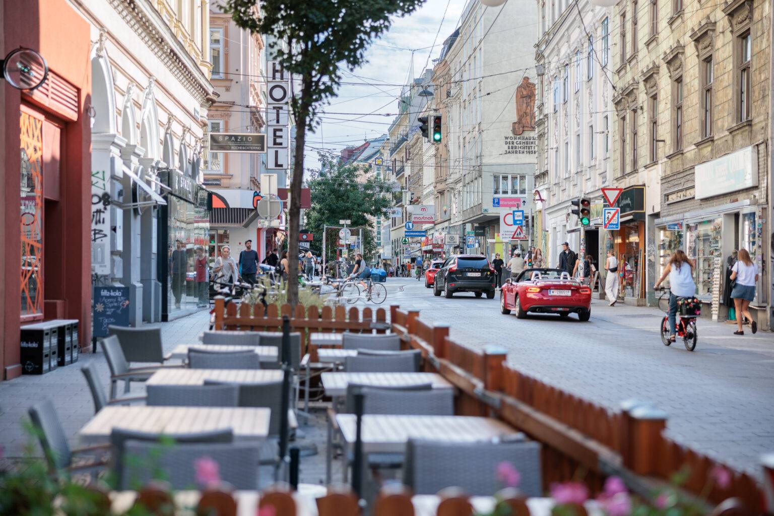 Neubaugasse mit Cafétischen im Vordergrund, Autos und Fußgänger im Hintergrund, historische Gebäude entlang der Straße.