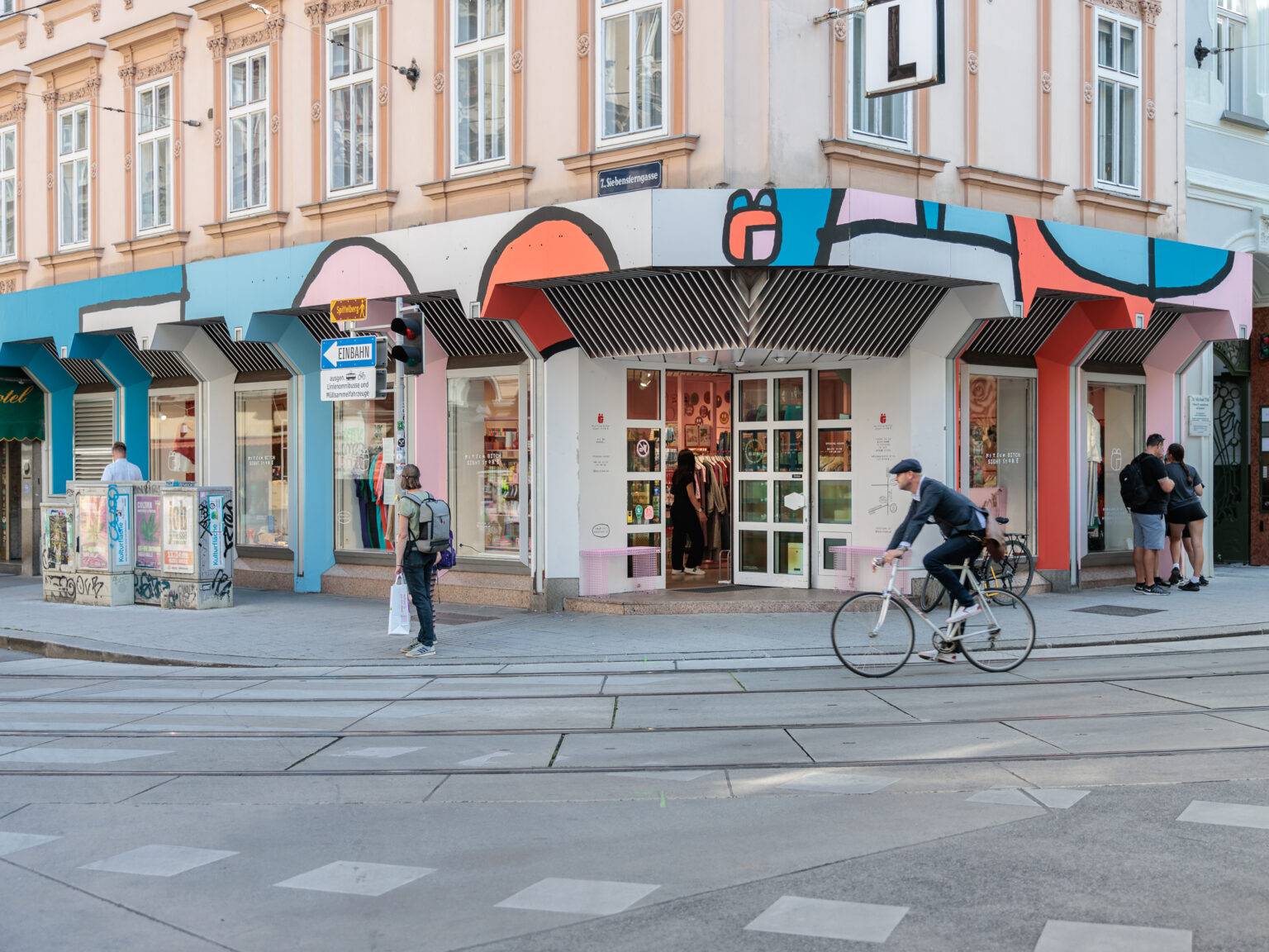 Bunte Fassade des Kitsch Bitch Store an einer Straßenkreuzung, mit Passanten und einem Radfahrer.