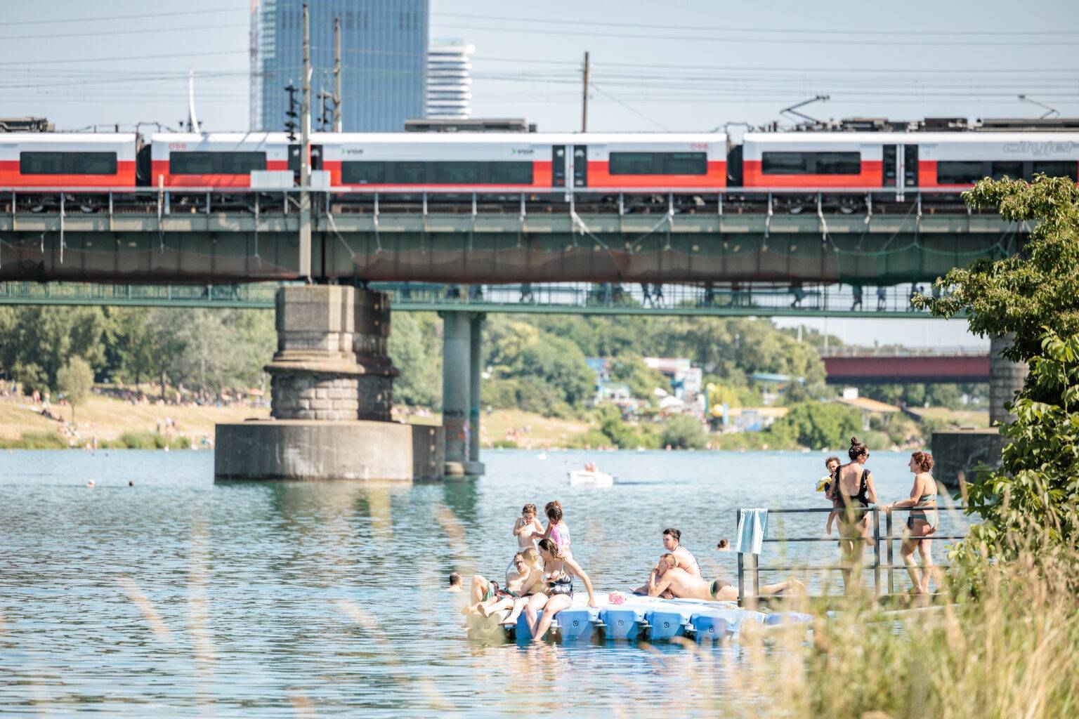 Menschen entspannen auf einem Floß im Wasser auf der neuen Donau, während ein Zug über eine Brücke fährt.