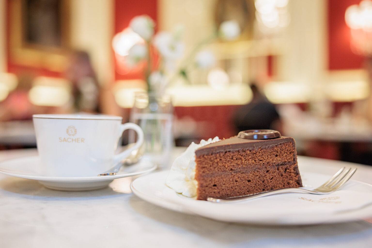 Tasse Kaffee im Kaffeehaus Sacher neben einem Stück Sachertorte auf einem Teller, mit einer Vase Blumen im Hintergrund.