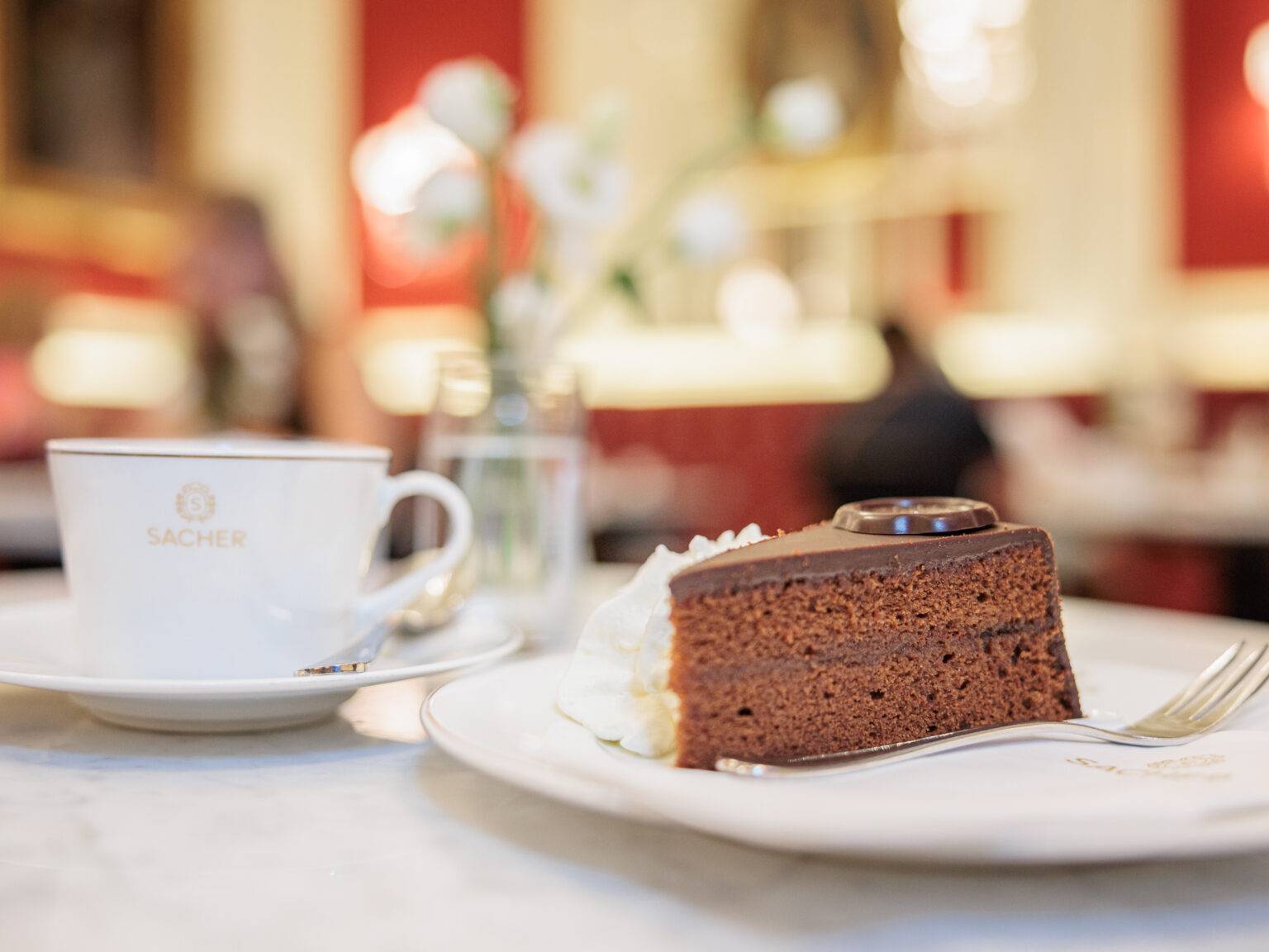 Tasse Kaffee im Kaffeehaus Sacher neben einem Stück Sachertorte auf einem Teller, mit einer Vase Blumen im Hintergrund.