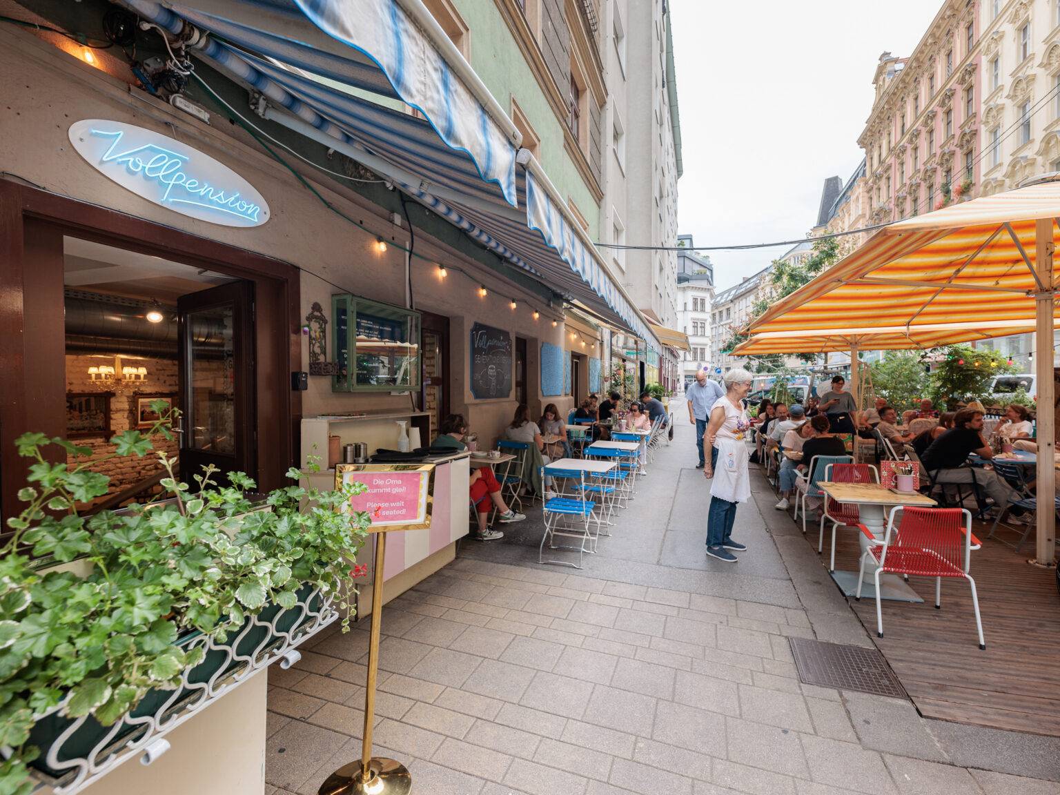 Belebter Außensitzbereich des Café Vollpension mit Tischen im Freien und bunten Sonnenschirmen.