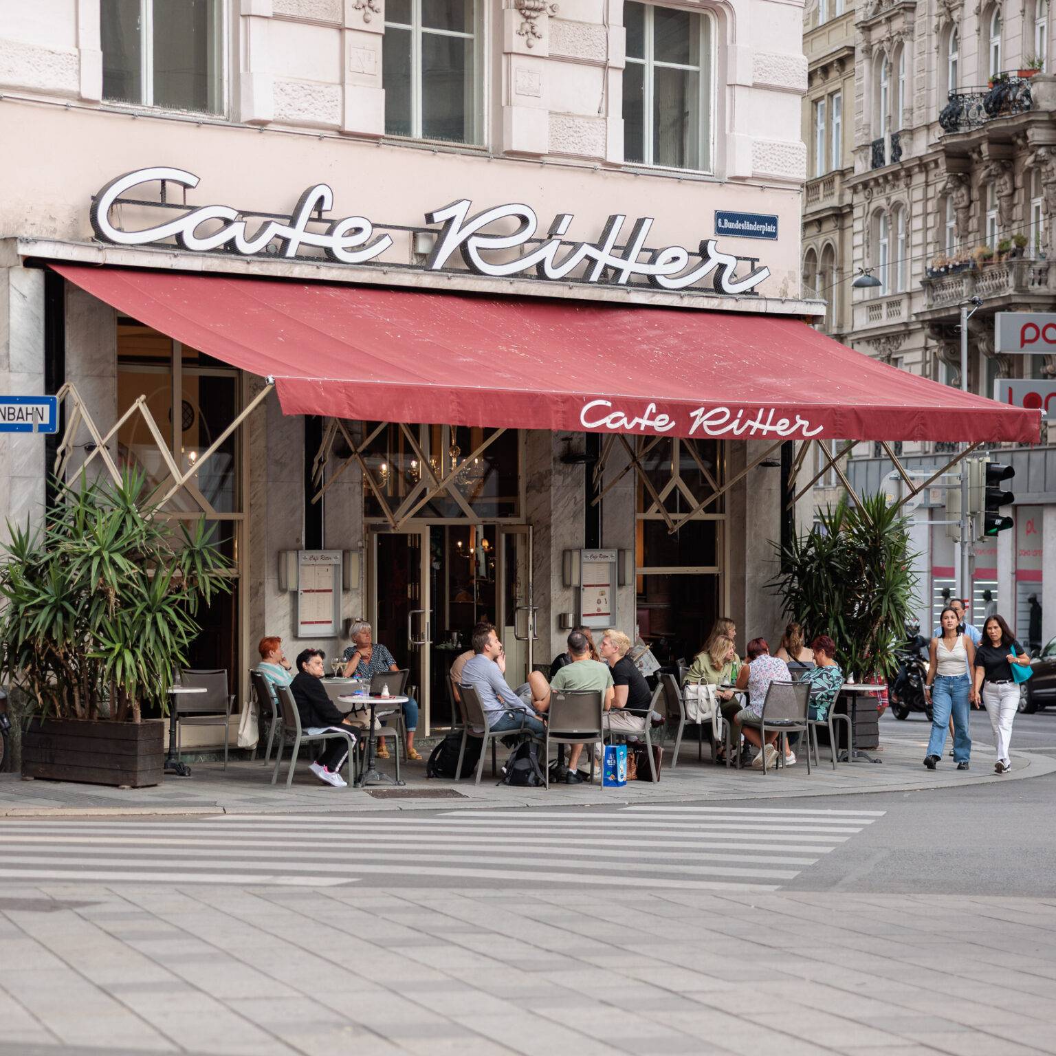 Café Ritter mit roter Markise, Tischen und Gästen auf der Terrasse in einer städtischen Umgebung.
