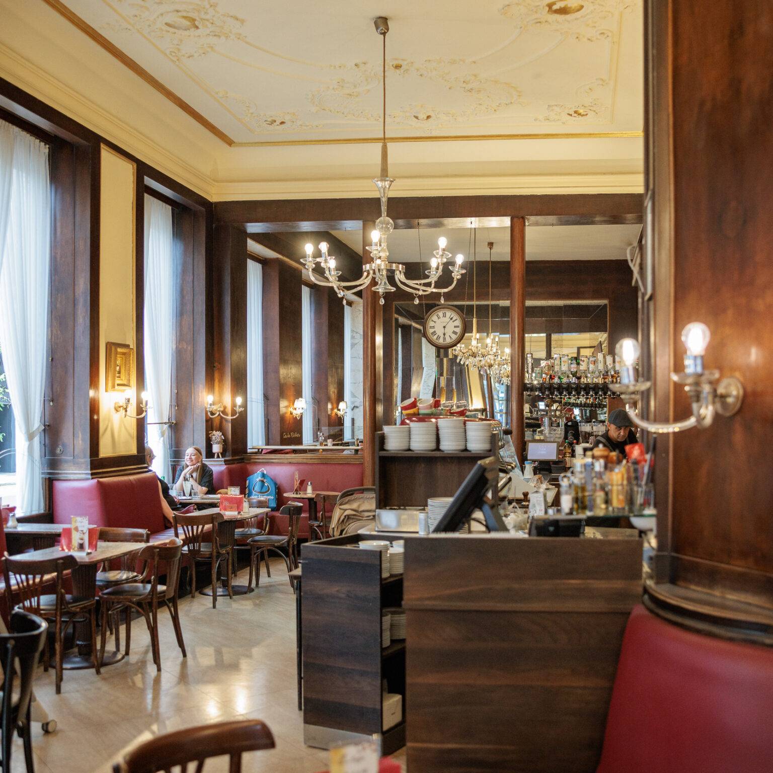 Gemütliches Café Ritter mit roten Polstermöbeln, einem Bartresen aus dunklem Holz und großen Fenstern, die viel Licht hereinlassen.