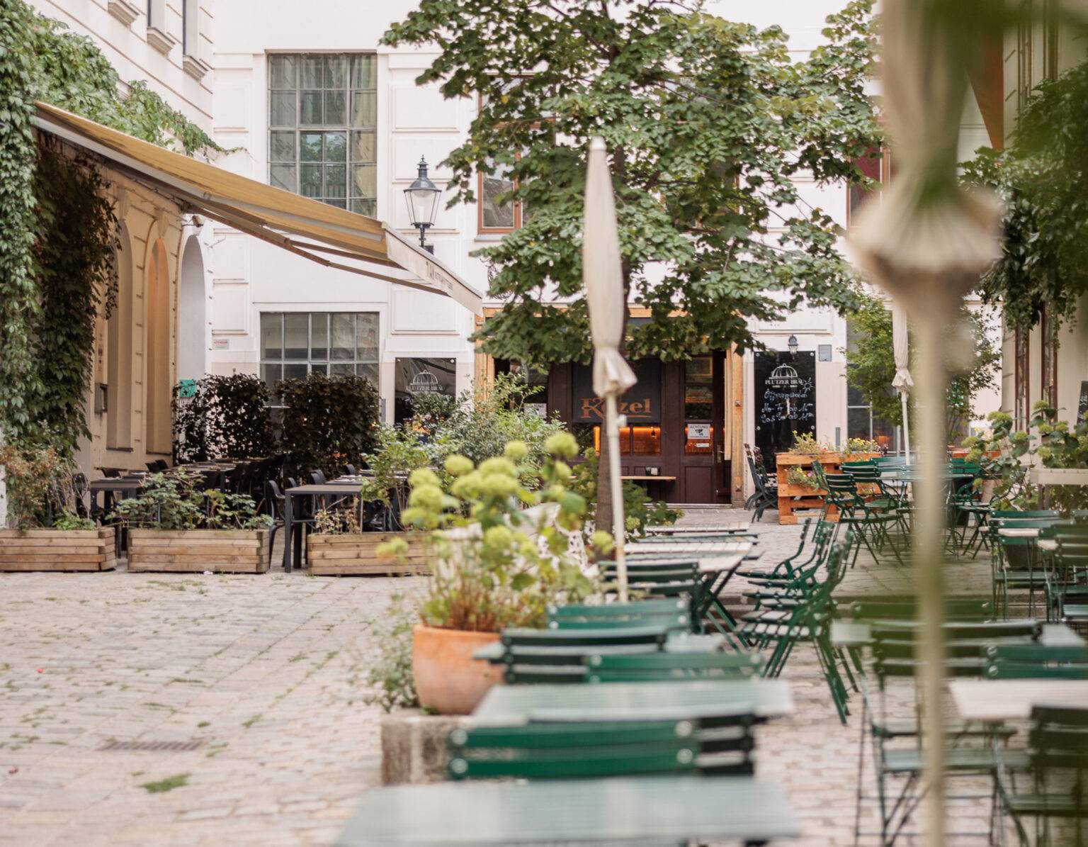 Gemütlicher Innenhof mit grünen Tischen, Pflanzen und einem kleinen Café im Hintergrund.