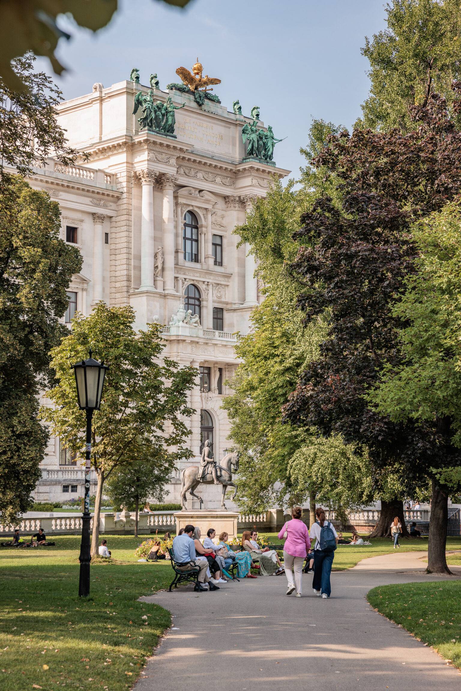Menschen sitzen auf Bänken im Burggarten, umgeben von Bäumen, mit der historischen Hofburg im Hintergrund.