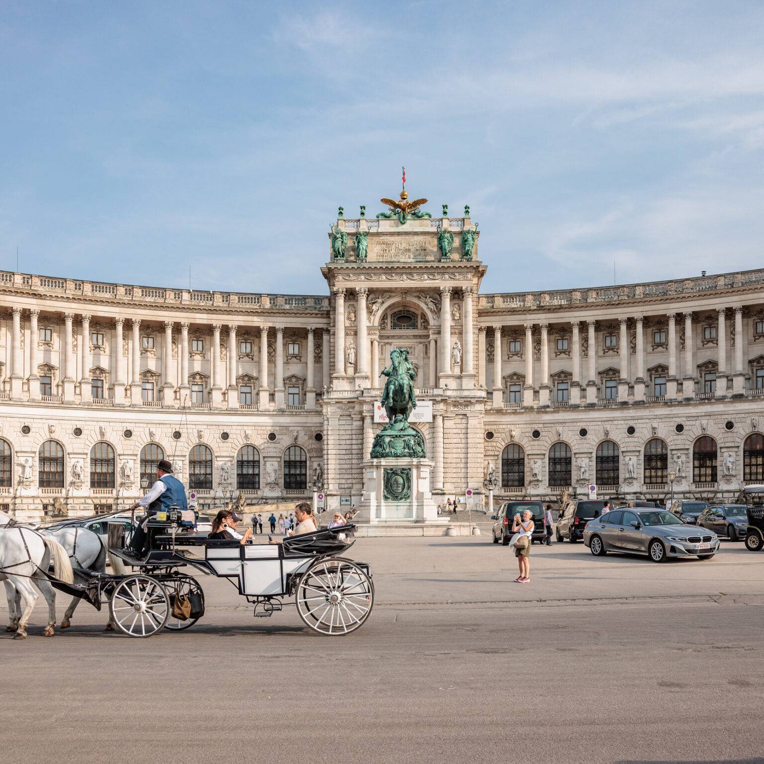 Pferdekutsche auf dem Heldenplatz vor der Hofburg mit Säulen und Statue, umgeben von Flaggen und Fahrzeugen.
