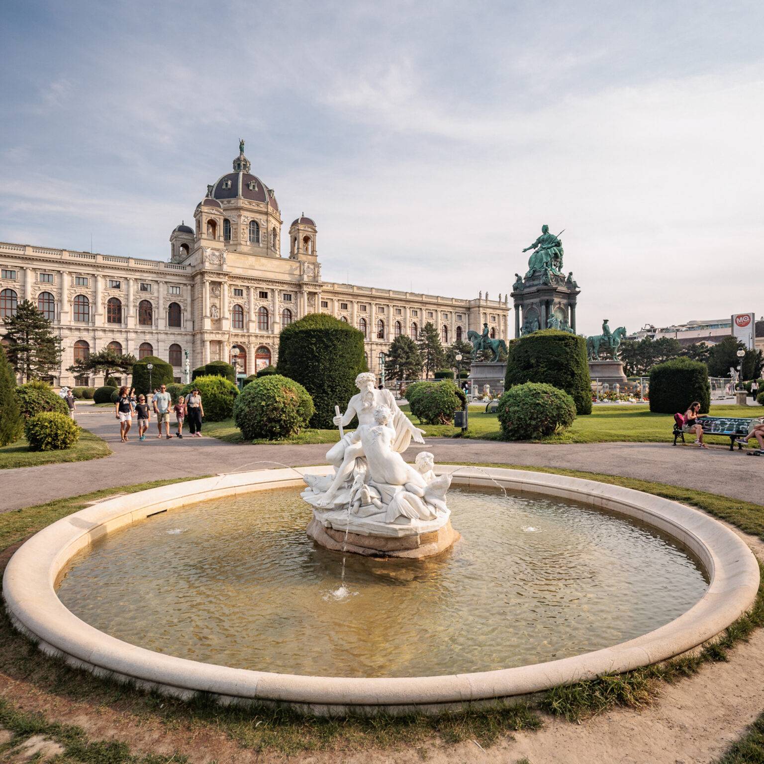 Brunnen mit Skulpturen in einem Park, umgeben von Büschen und dem KHM im Hintergrund.