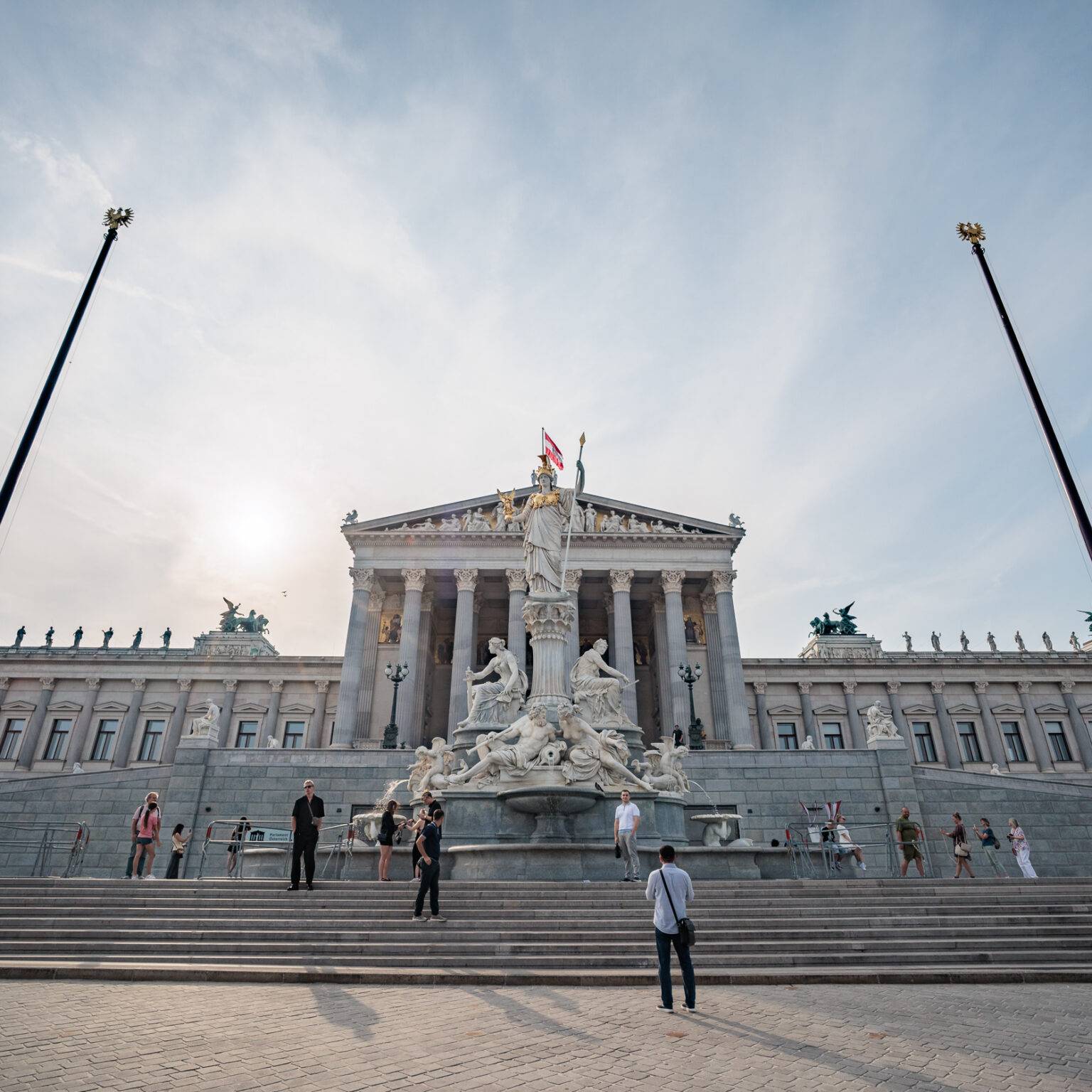 Historische Fassade des Parlaments mit Stufen, Statue im Vordergrund und Menschen, unter einem leicht bewölkten Himmel.